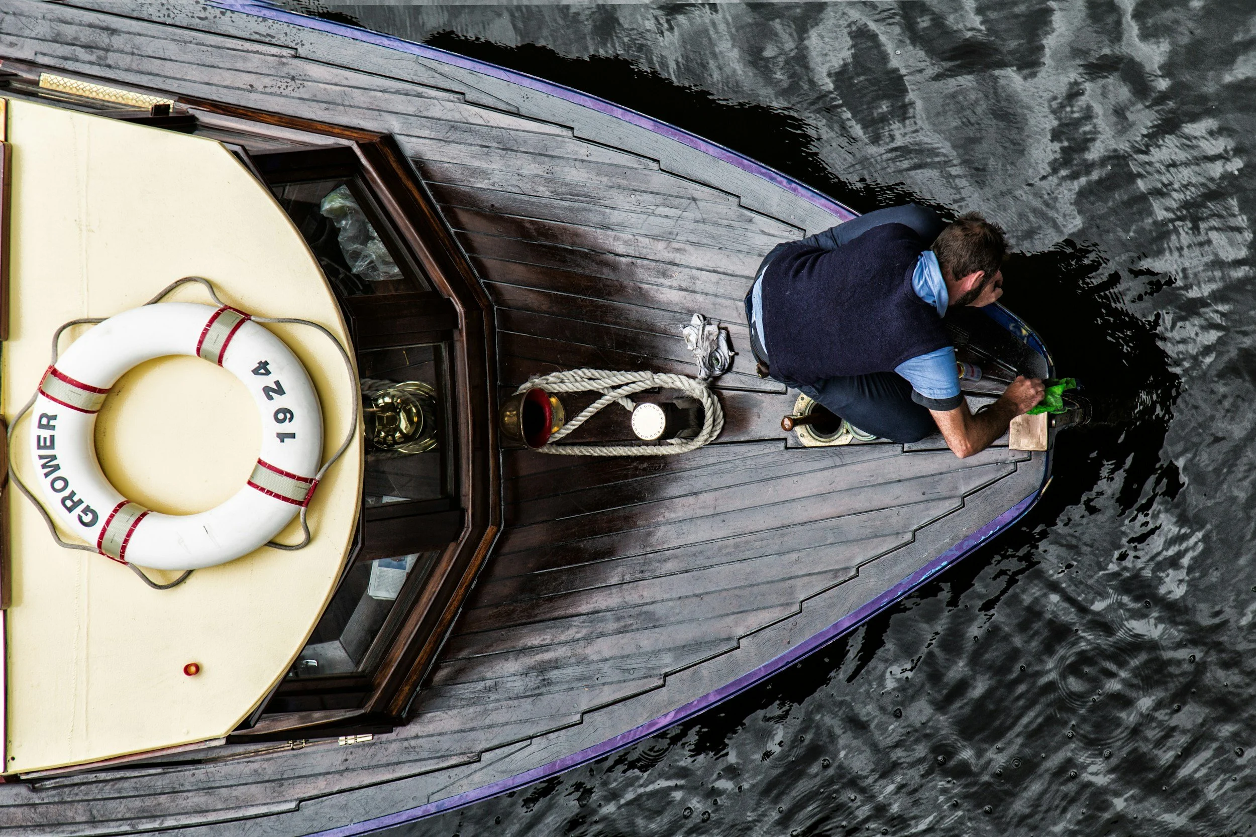 A man in a blue shirt and dark vest is on the deck of a wooden boat, looking into the water. The boat has a white lifebuoy with red stripes and text on it, along with a rope, a lantern, and some other equipment on the deck.