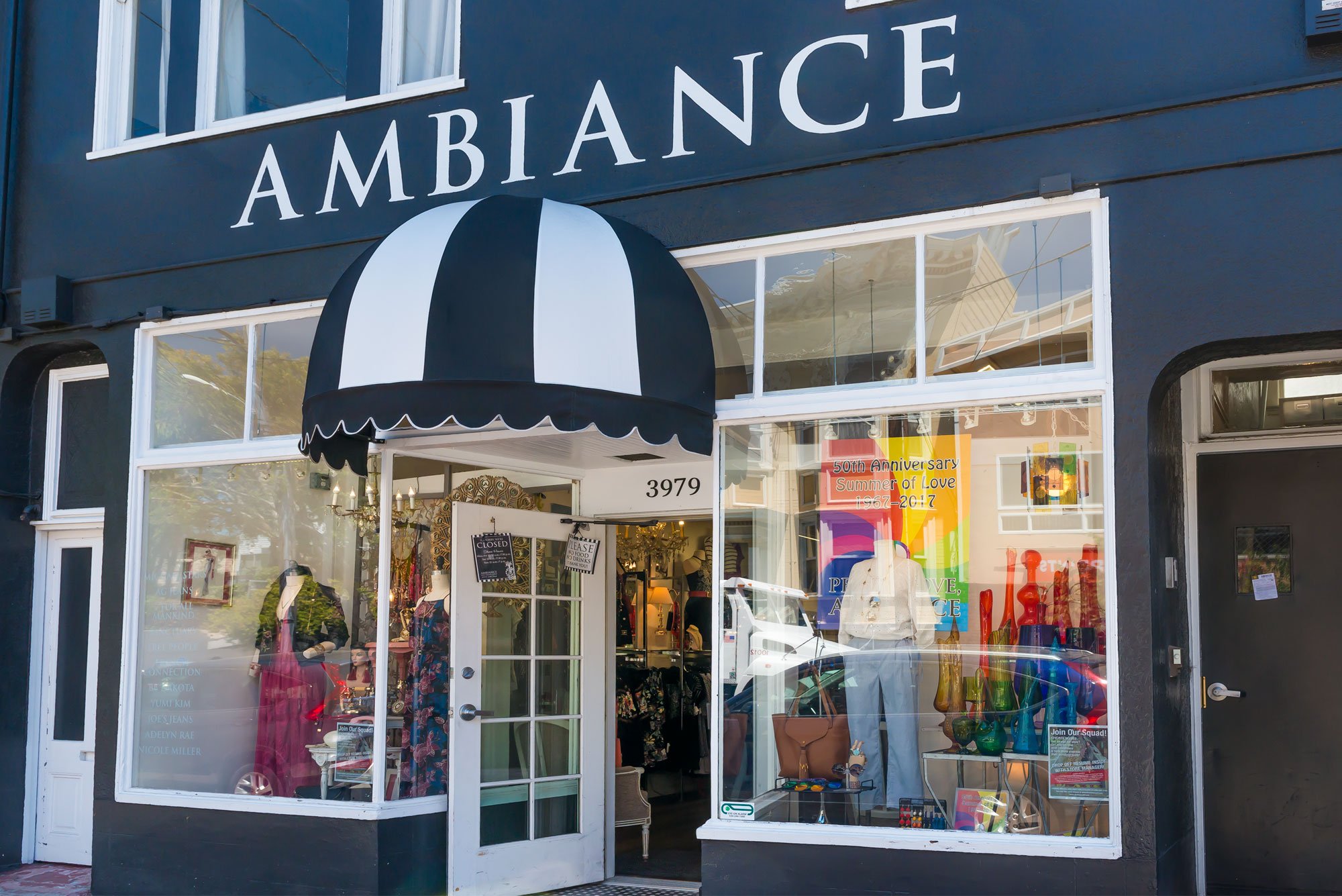 Storefront with a black and white striped awning, large glass windows displaying clothing and colorful glass vases, and a sign on the door indicating the store is closed.