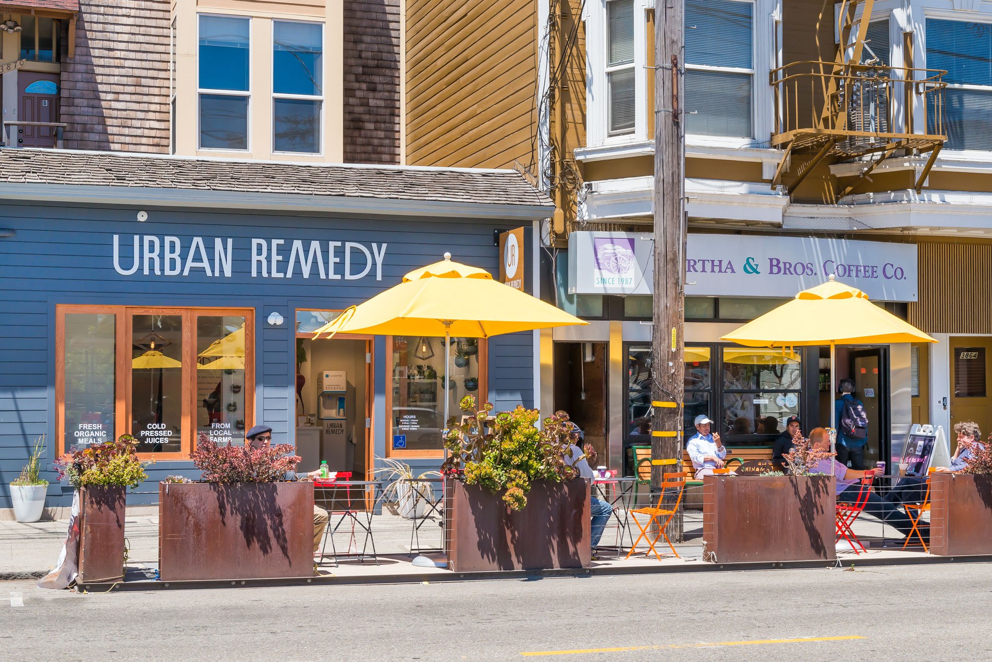 Street scene featuring a cafe with outdoor seating and yellow umbrellas. People sit at tables, some enjoying drinks. The storefronts include 'Urban Remedy' and 'Martha & Bros. Coffee Co.' with vibrant building facades in the background.