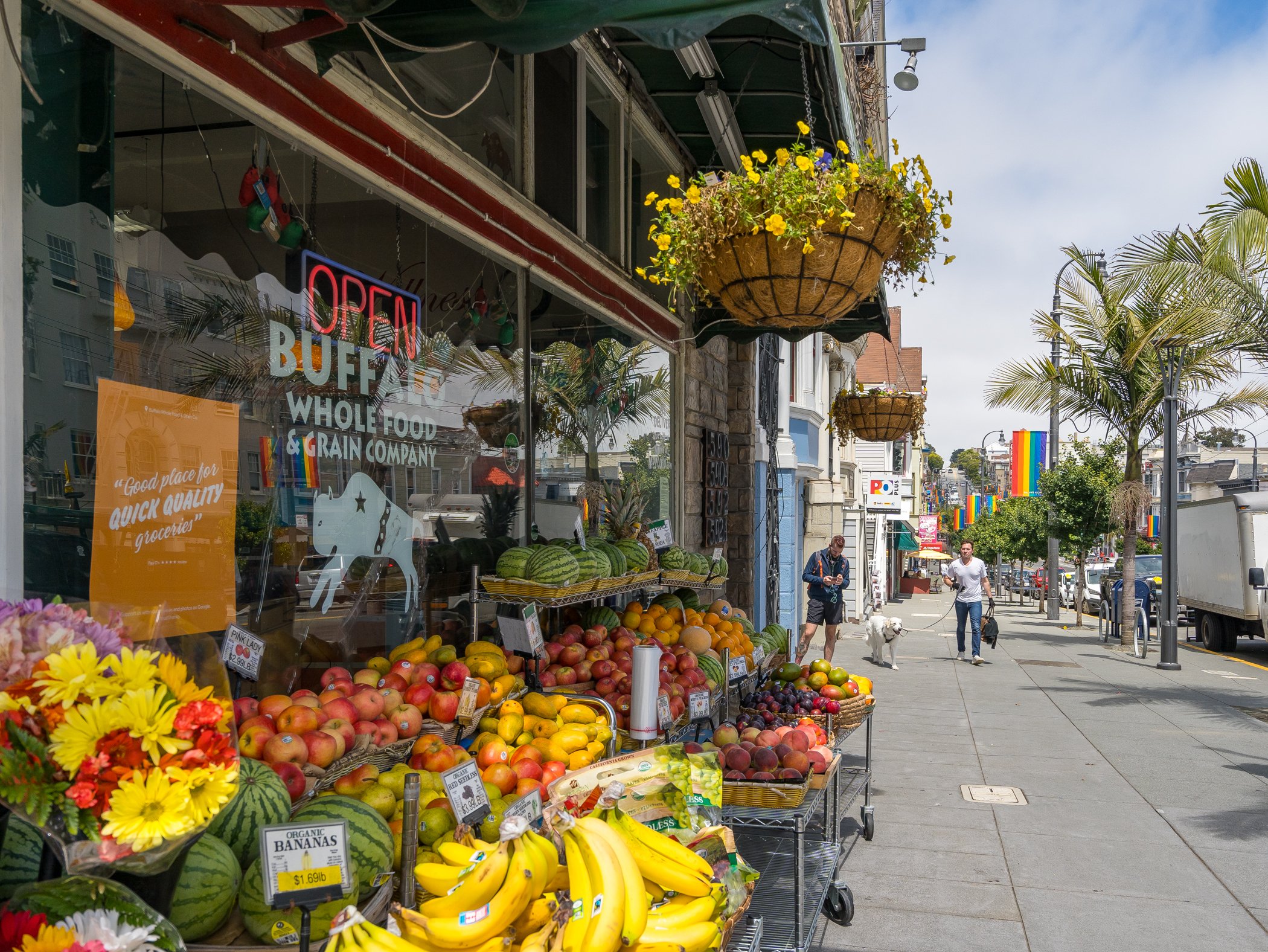 Fruit stand outside a store with watermelons, bananas, apples, and other fresh fruits, on a city sidewalk with pedestrians walking, trees, and rainbow pride flags