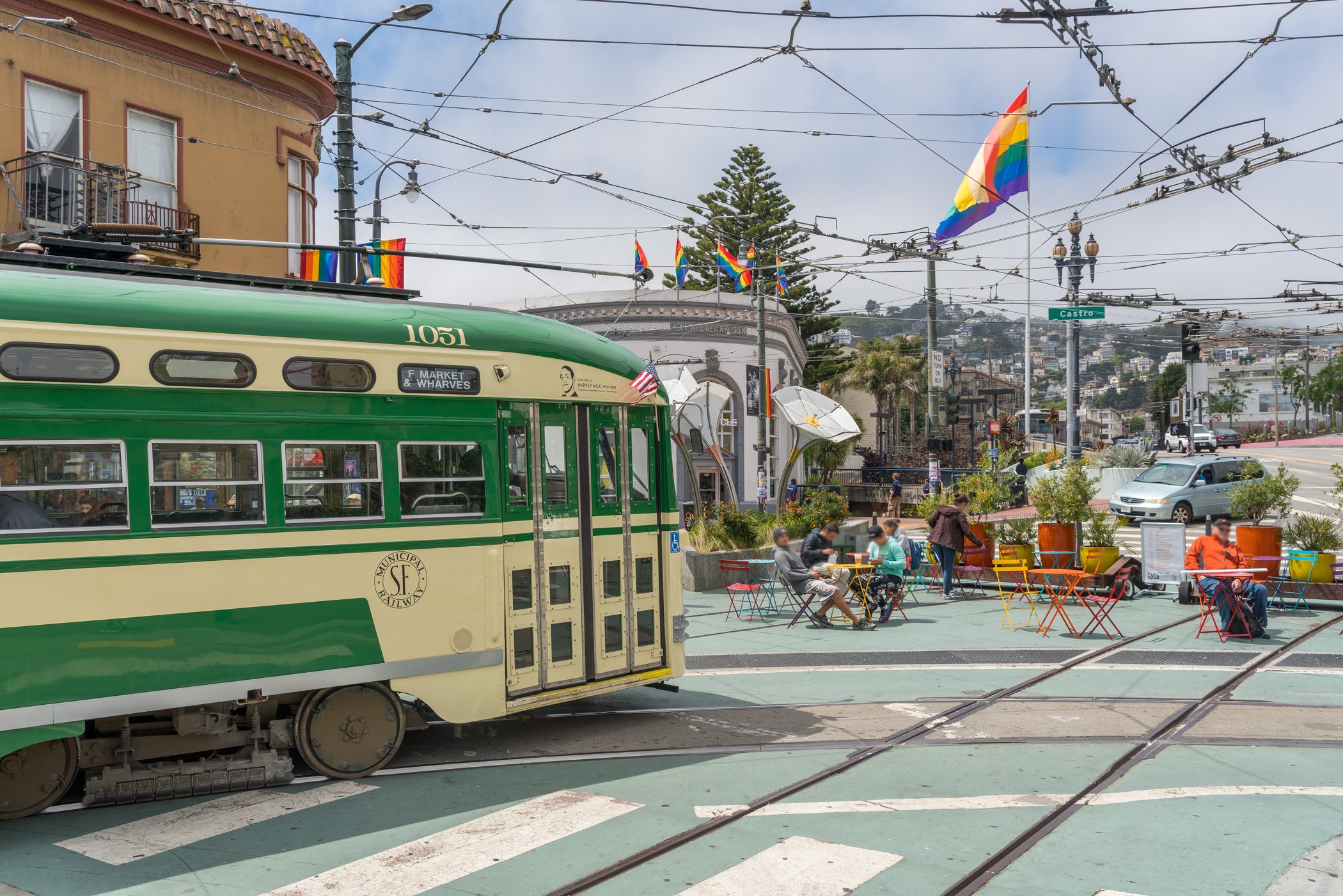 A green and cream municipal streetcar with the sign 'F Market & Wharves' on a city street with overhead trolley wires, Rainbow flags, a small outdoor seating area with people, and a building with a balcony, trees, and hills in the background.