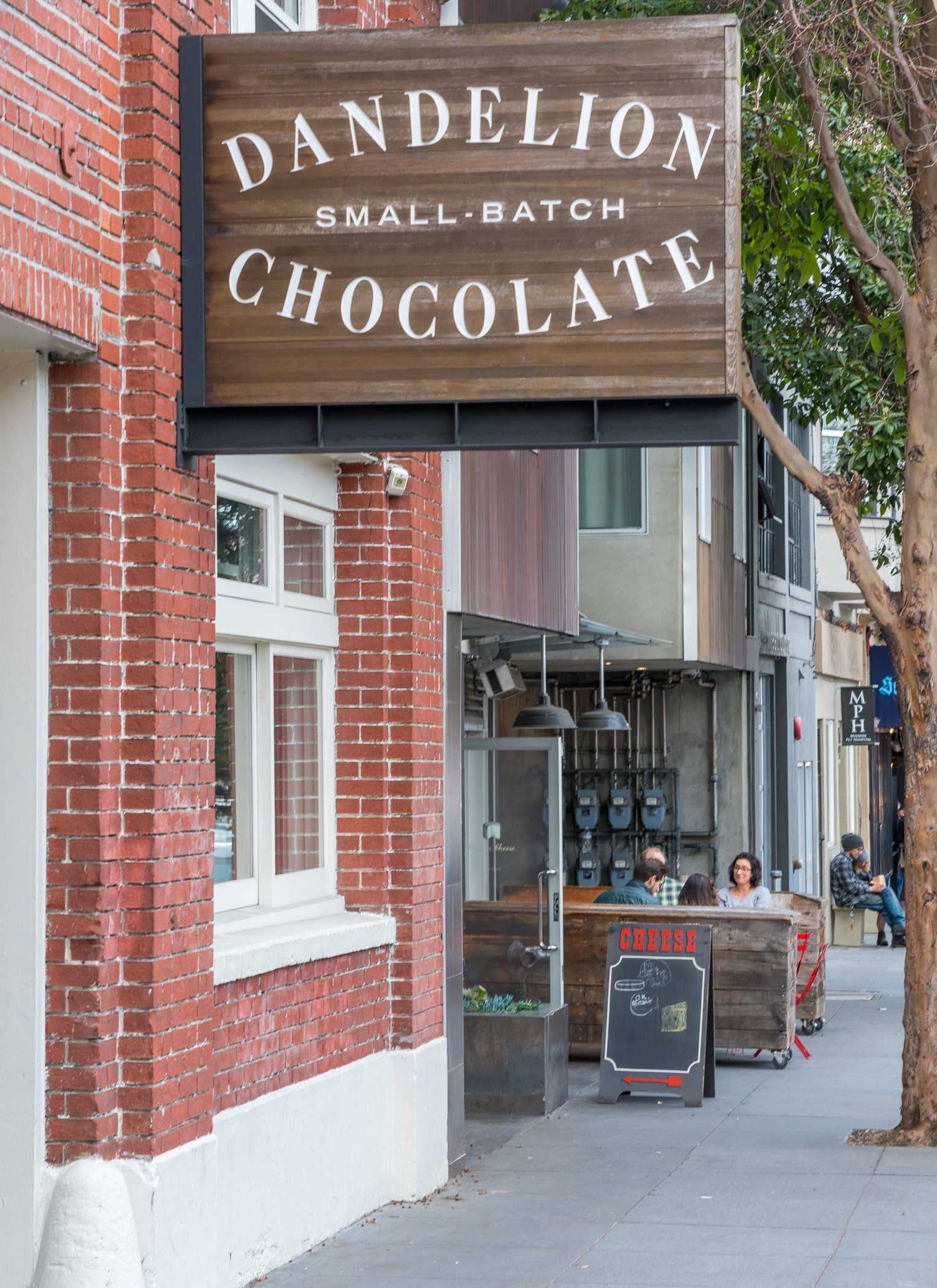 Street view showing a small retail storefront with a wooden sign that reads ''Dandelion Small-Batch Chocolate'' and a chalkboard sign that advertises cheese, with some people sitting at an outdoor table of a nearby cafe, and red-brick and modern buil