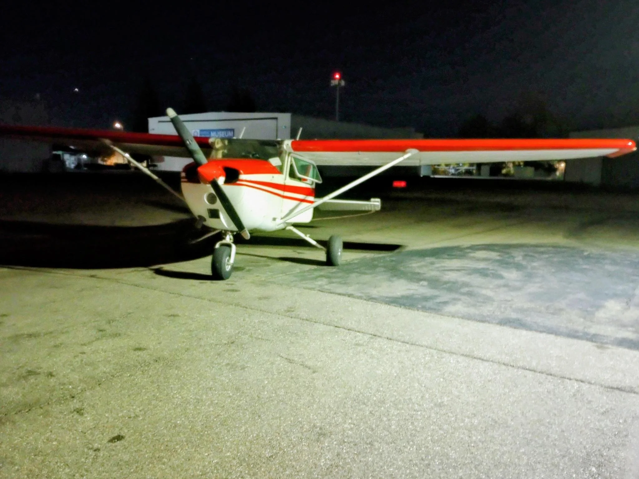 Small single-engine airplane with white, red, and green colors parked on an airport tarmac at night, illuminated by bright ground lights.