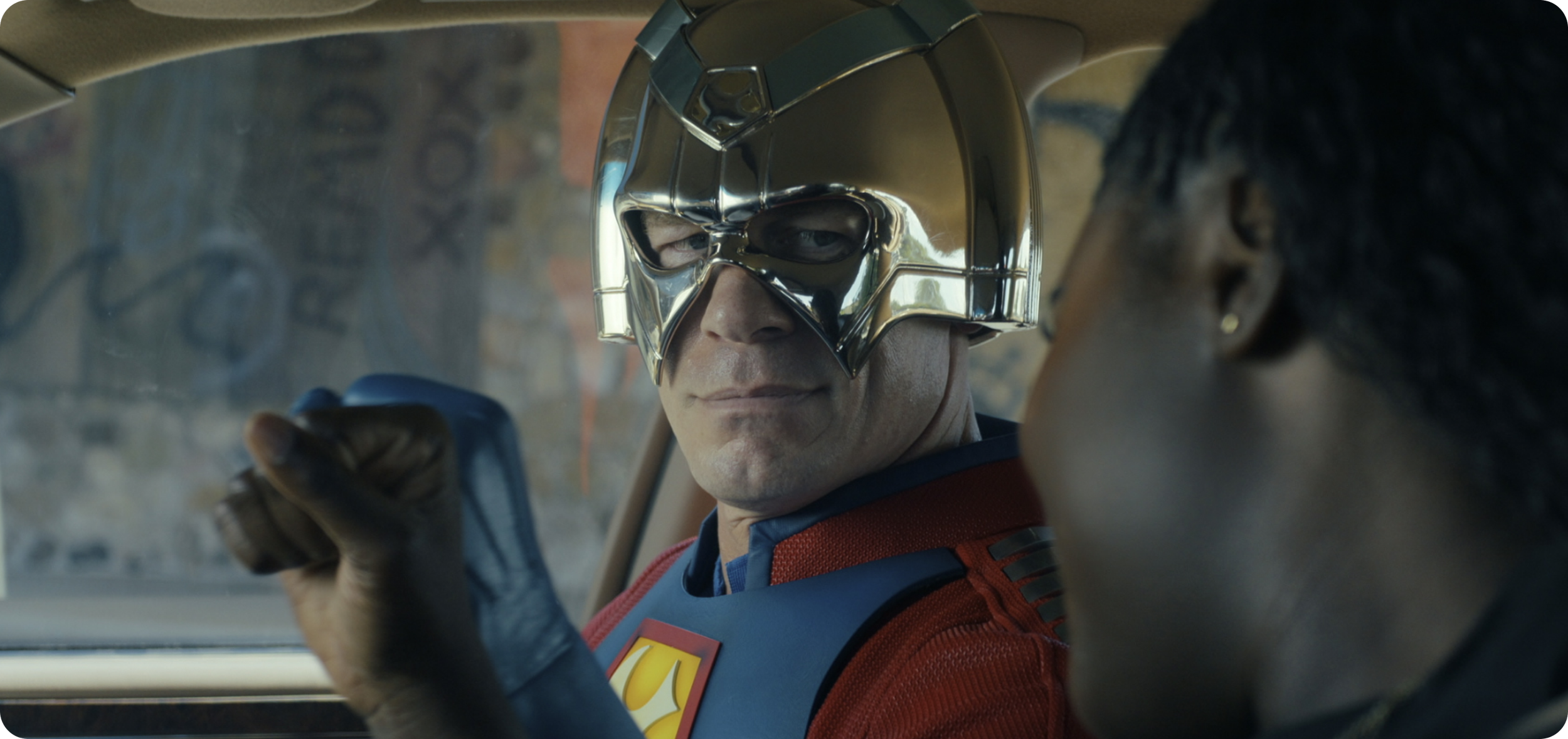 A man wearing a shiny, metallic mask and superhero costume sitting in a car, looking at a woman with braided hair with a serious expression.