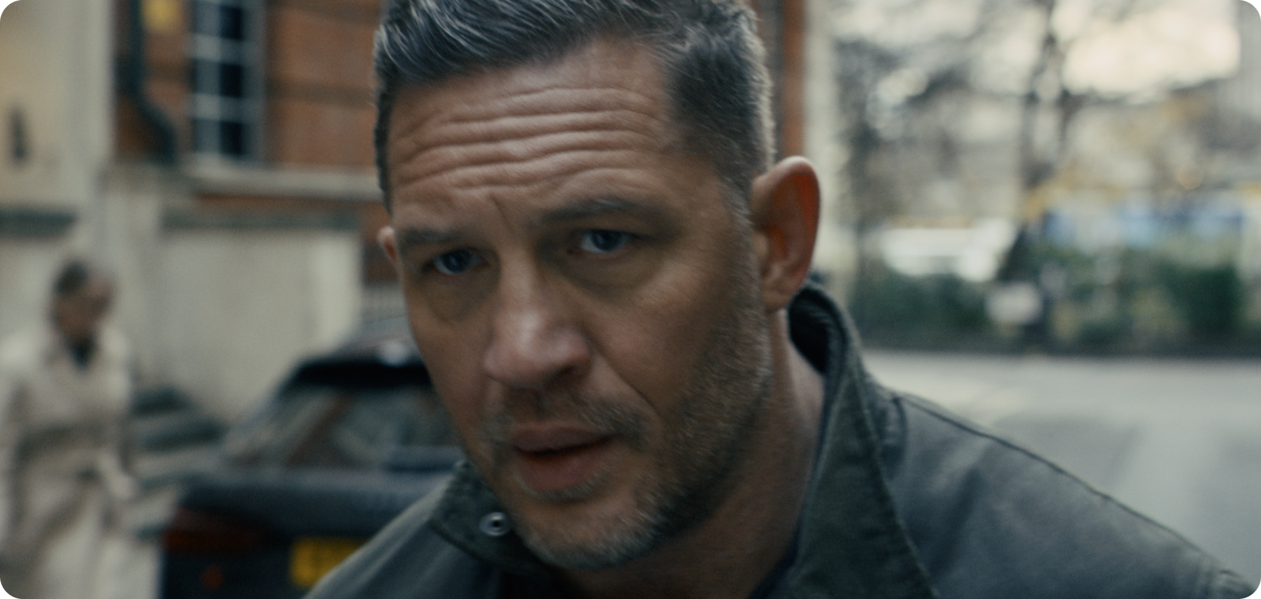 Close-up of a man with light skin, short gray hair, and blue eyes, wearing a dark leather jacket, with an intense expression on his face.