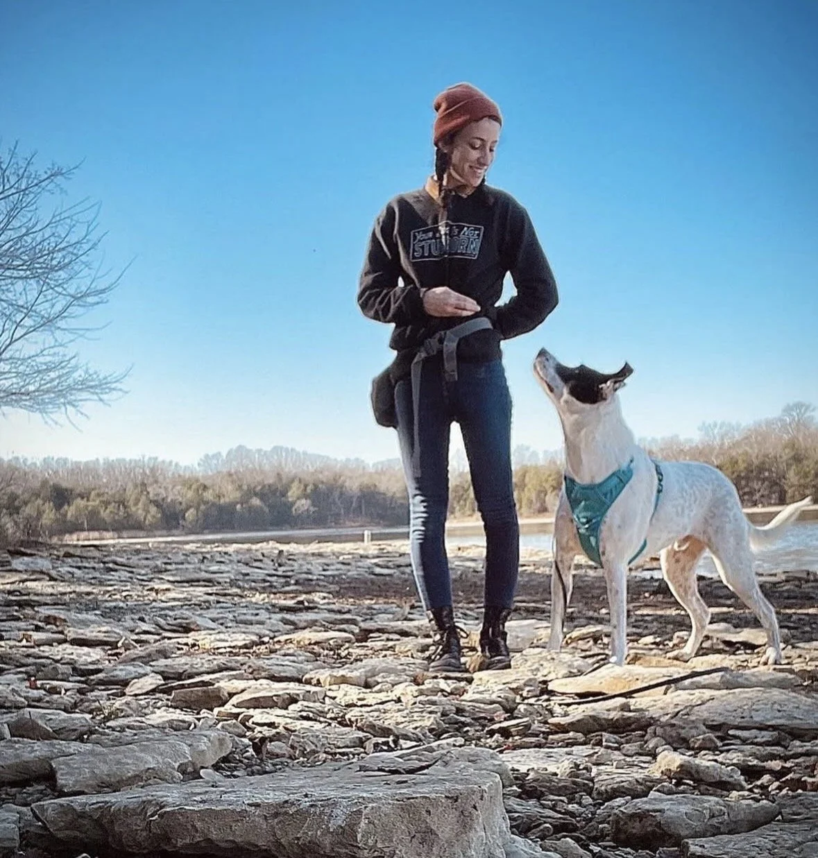 A woman and a dog standing on a rocky shoreline near a body of water with trees in the background under a clear blue sky.