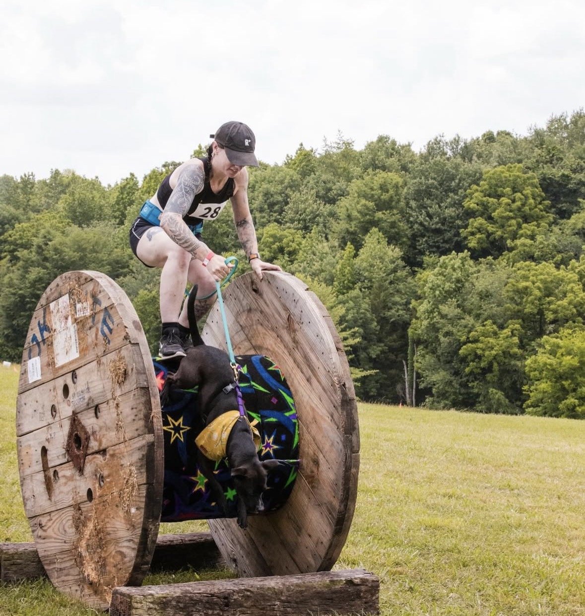Woman with tattoos and a black cap navigating a large wooden spool with her dog inside, during an outdoor event on a grassy field with trees in the background.