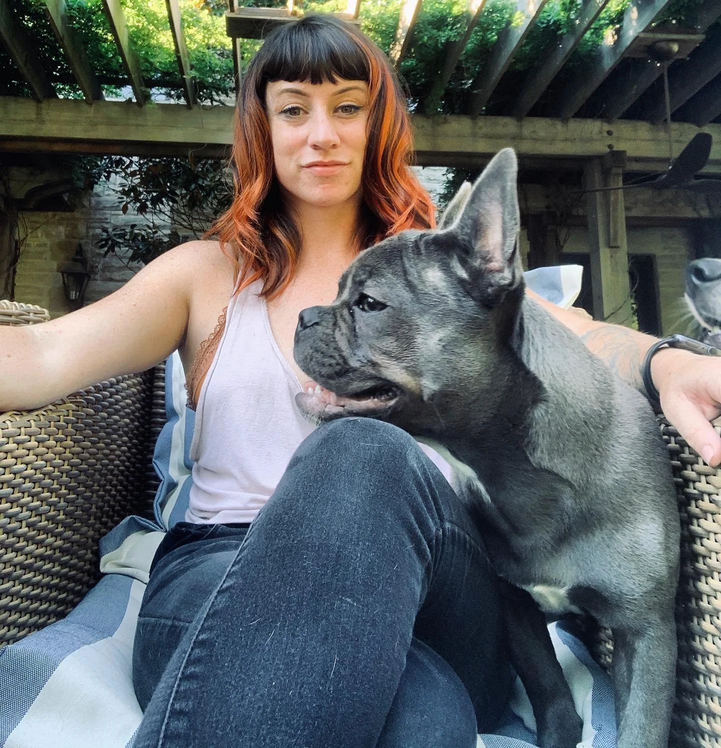 A woman with red hair sitting outdoors on a wicker couch, holding a young dog with a brindle coat and large ears, in a backyard with greenery and a wooden structure in the background.