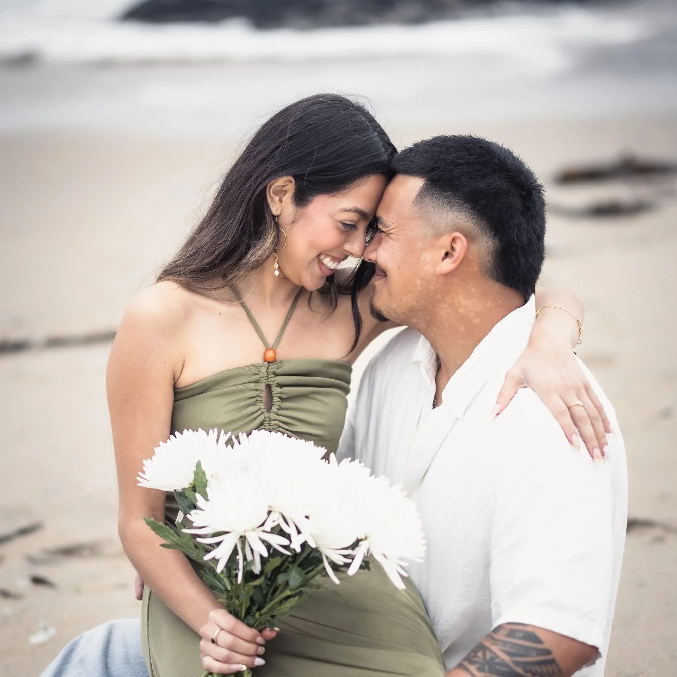 Couple smiles at each other as they sit on the beach holding flowers.