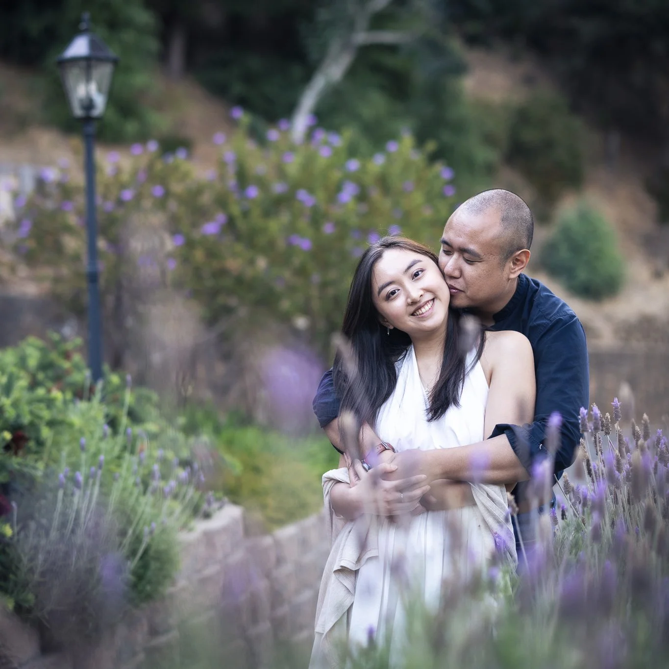 Groom-to-be kisses his fiancee's neck, surrounded by lavender during engagement shoot.