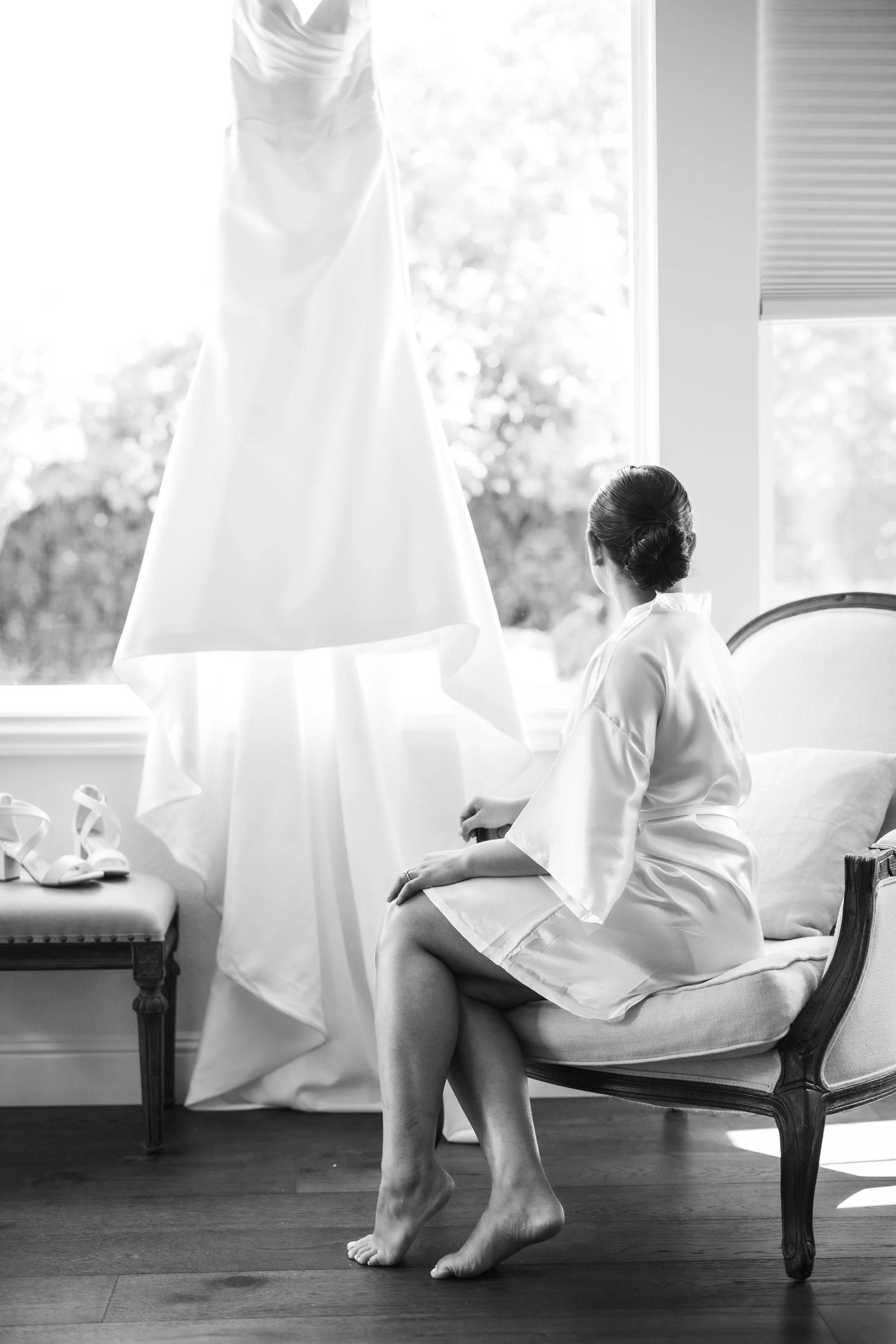 Bride sits in her robe gazing at her wedding dress.