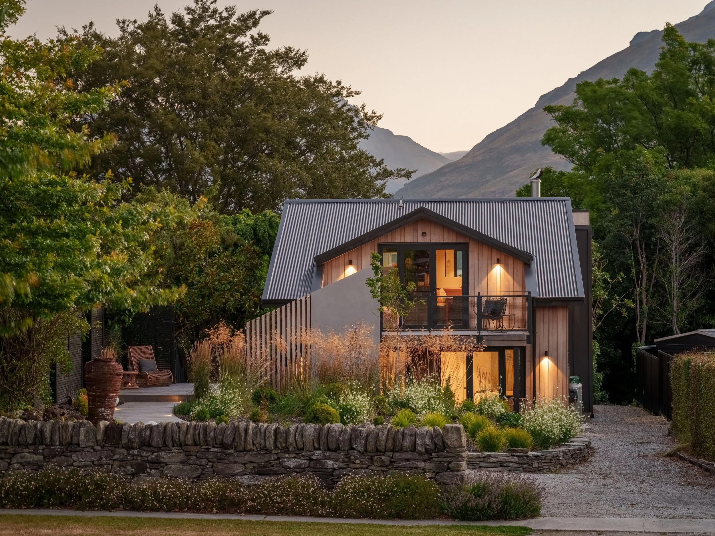 A modern house with a steep metal roof and large windows, surrounded by lush trees and a garden with plants and flowers, mountain landscape in the background at dusk.