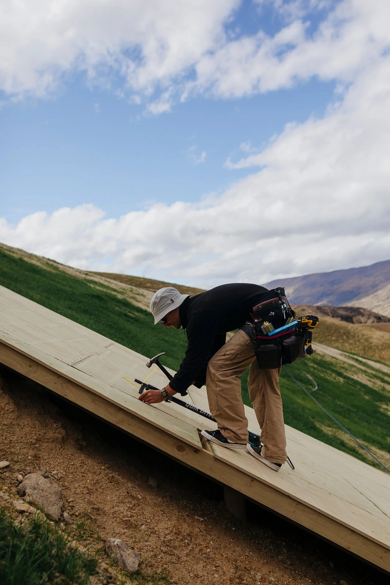 A man working on the construction of a wooden sloped roof outdoors, wearing a white hat and black hoodie, with tools and equipment hanging from a belt
