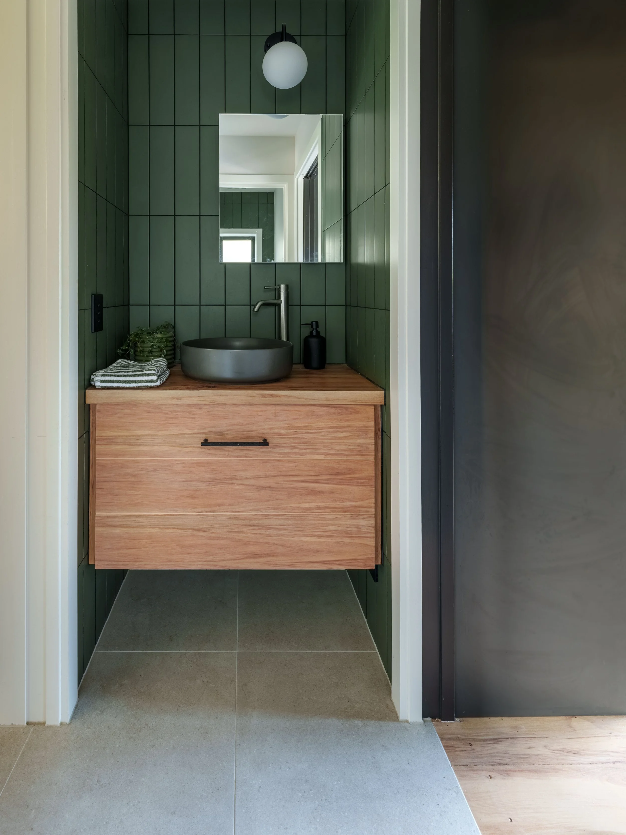 A modern bathroom vanity with a round, dark gray vessel sink, a silver faucet, a black soap dispenser, a folded gray and white striped towel, a small green plant, and a mirror on a green tiled wall. The vanity is made of light wood, and there is a sp