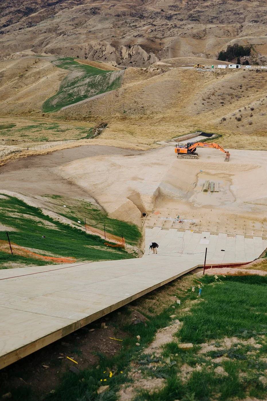 Construction site on a hillside with excavator digging and workers assembling a section of pavement.