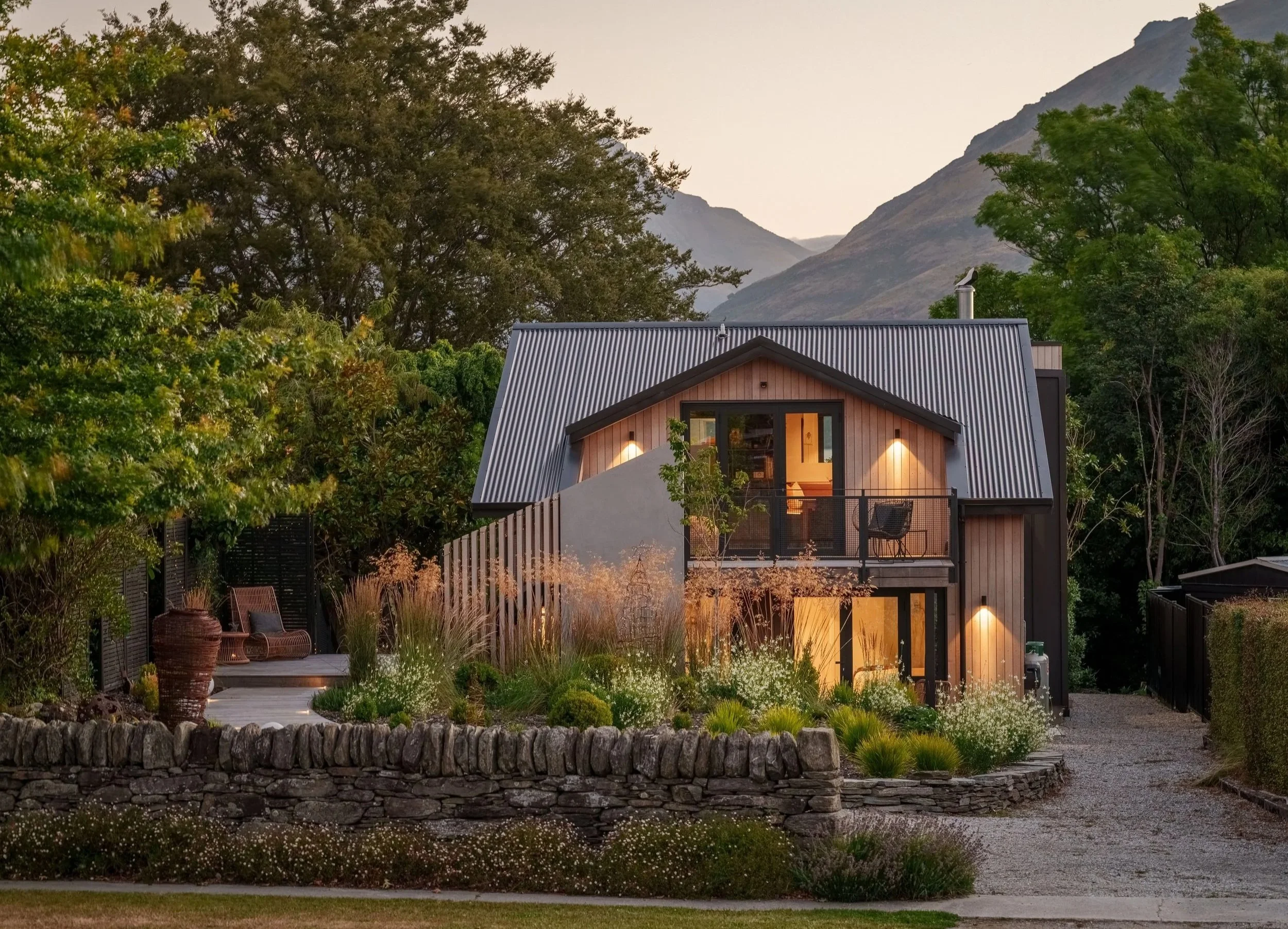 Modern house with metal roof, large windows, and outdoor deck surrounded by trees and garden, mountainous landscape in the background during dusk.