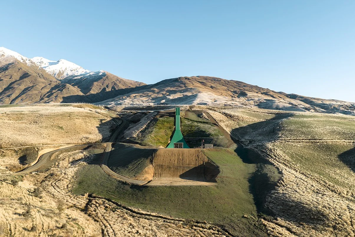 A ski jump built on a grassy hillside with mountainous terrain and a clear blue sky in the background.
