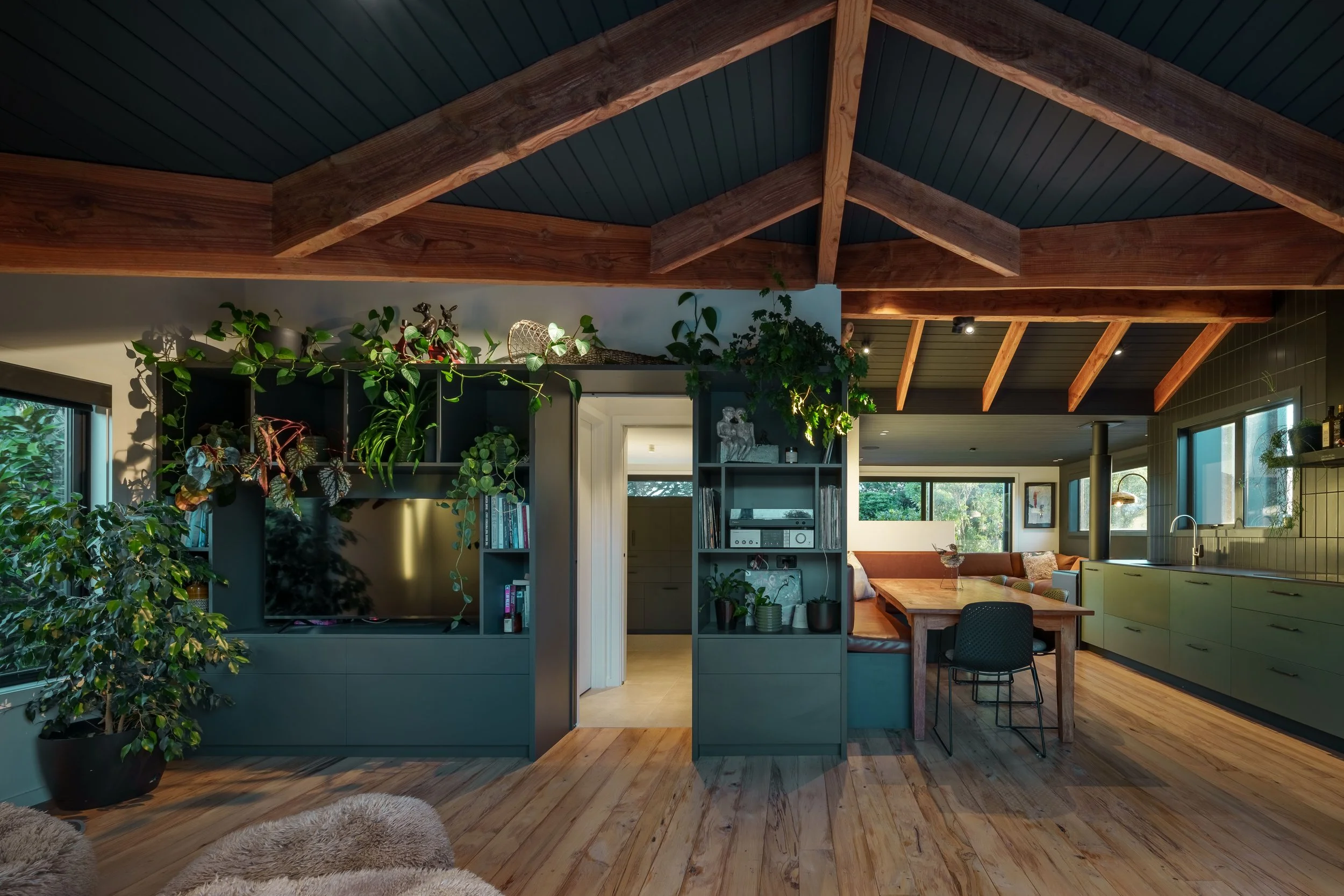 Interior view of a living room with a vaulted ceiling featuring wooden beams and a dark ceiling panel. The room is decorated with numerous green houseplants on shelves and stands. There is a large flat-screen TV, books, and decorative objects. A door