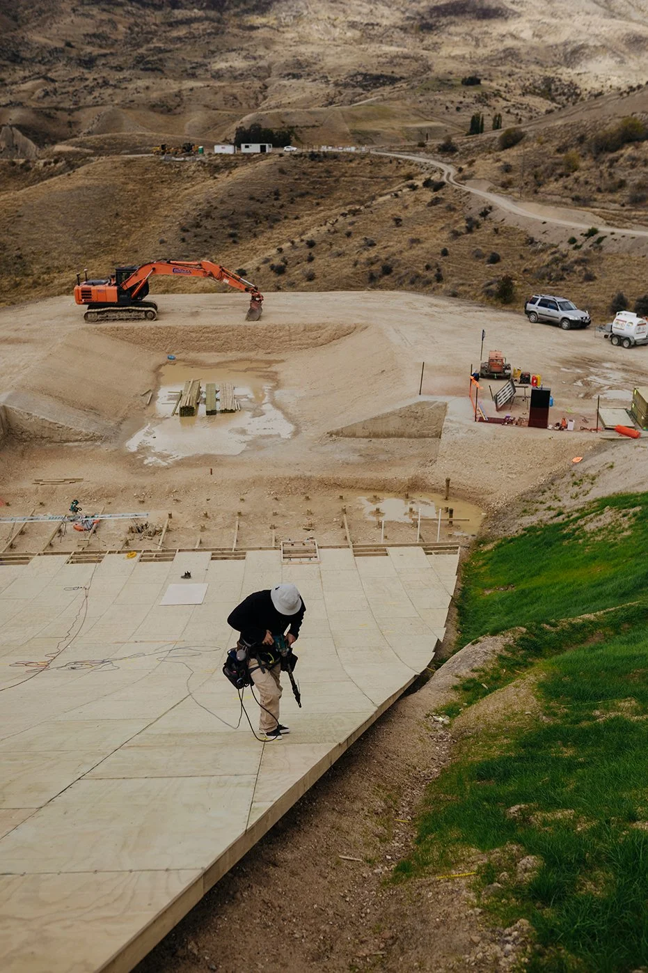A construction worker working on a large wooden platform at a construction site in a hilly, semi-rural area with a dirt road and a few parked vehicles in the background.