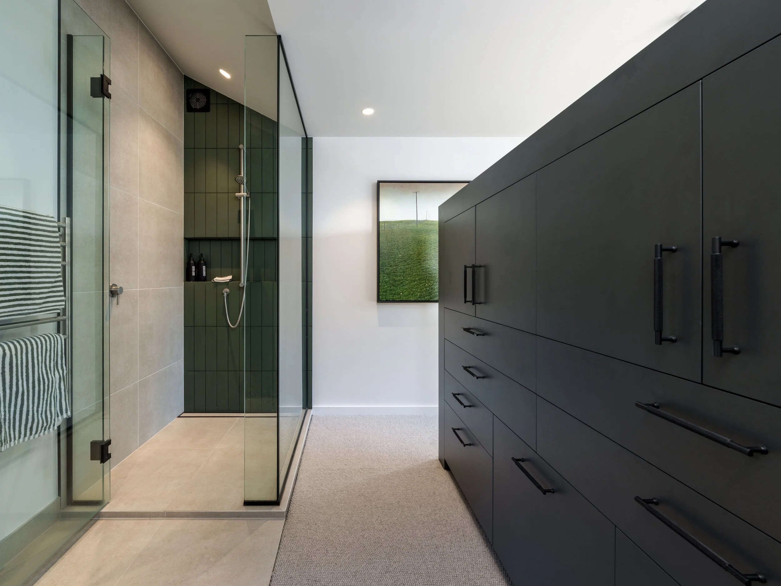 Modern bathroom with walk-in shower, dark cabinetry, and framed artwork of green field and sky.
