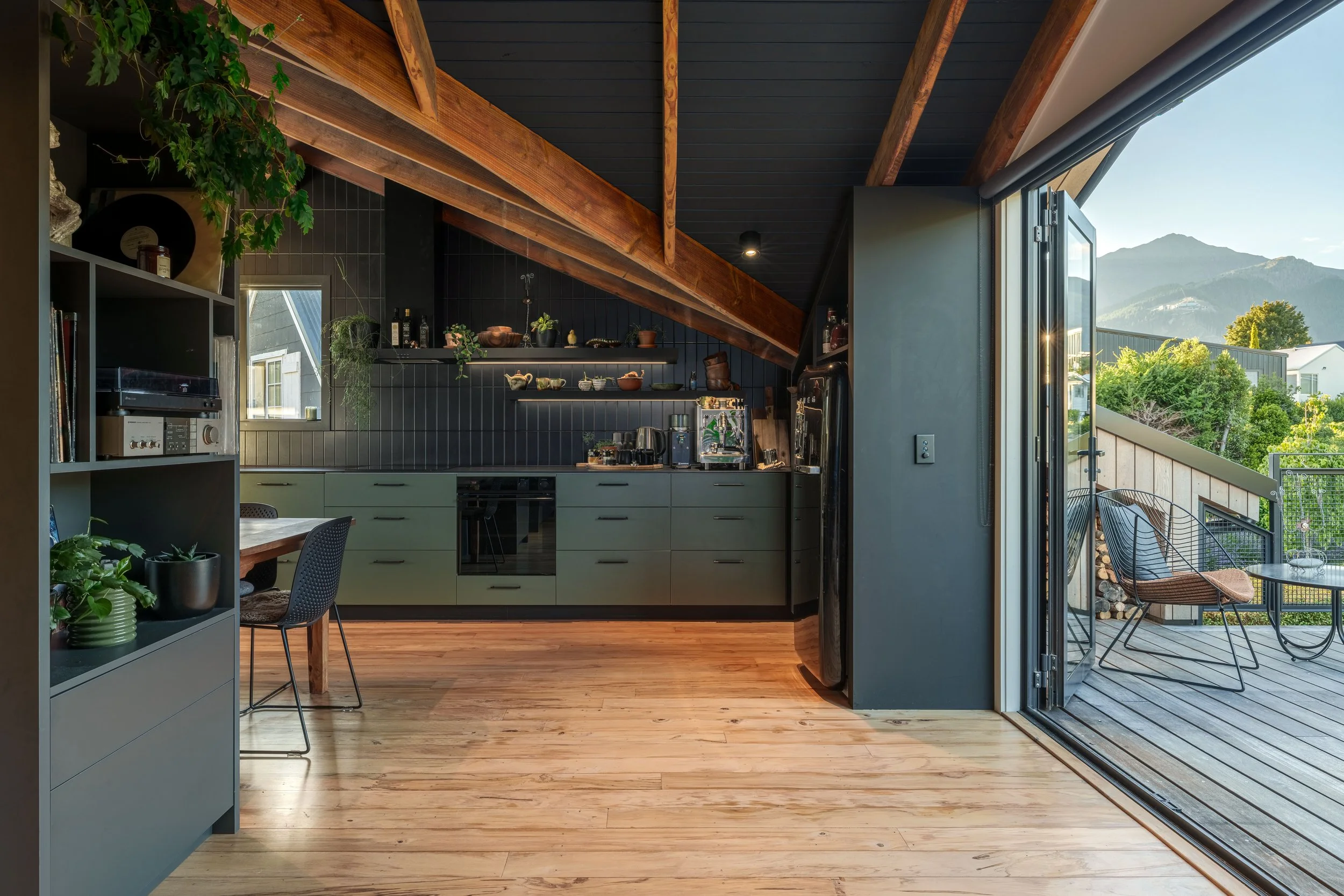 Modern kitchen with black ceiling and wood beams, gray cabinets, and an open sliding glass door leading to a balcony with outdoor chairs, mountains, and blue sky in the background.