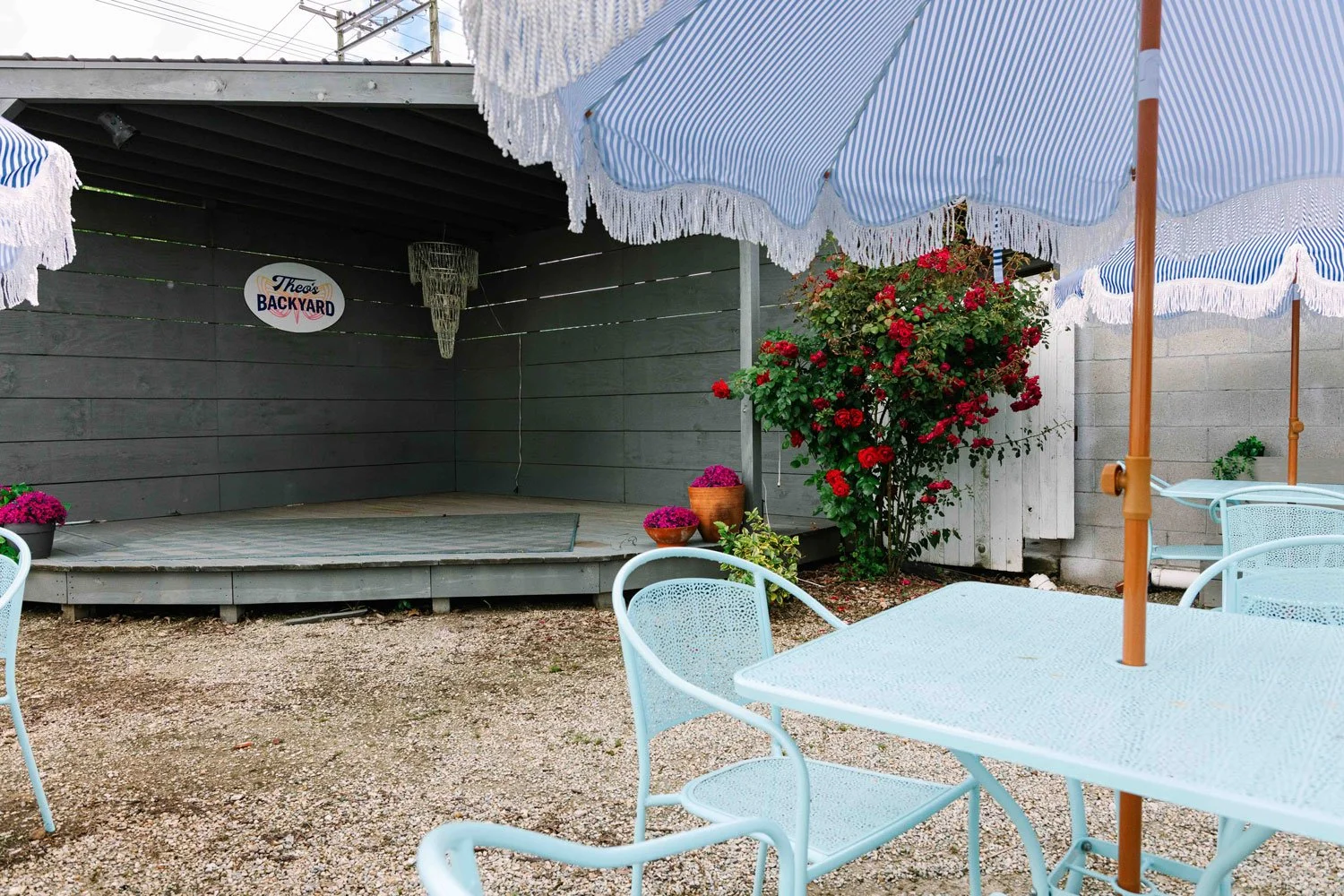 Outdoor backyard patio with blue and white striped umbrellas, white metal chairs, a small stage, potted flowers, and red blooming plants against a wooden and concrete fence.