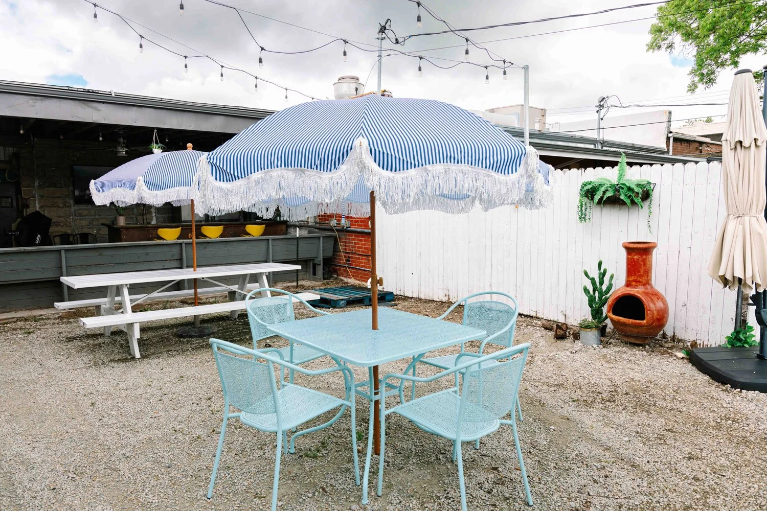 An outdoor patio area with light blue metal furniture including a table and four chairs under a blue and white striped umbrella with fringe. There is a white wooden picnic table nearby, a white fence with plants and a large terracotta chiminea, and s