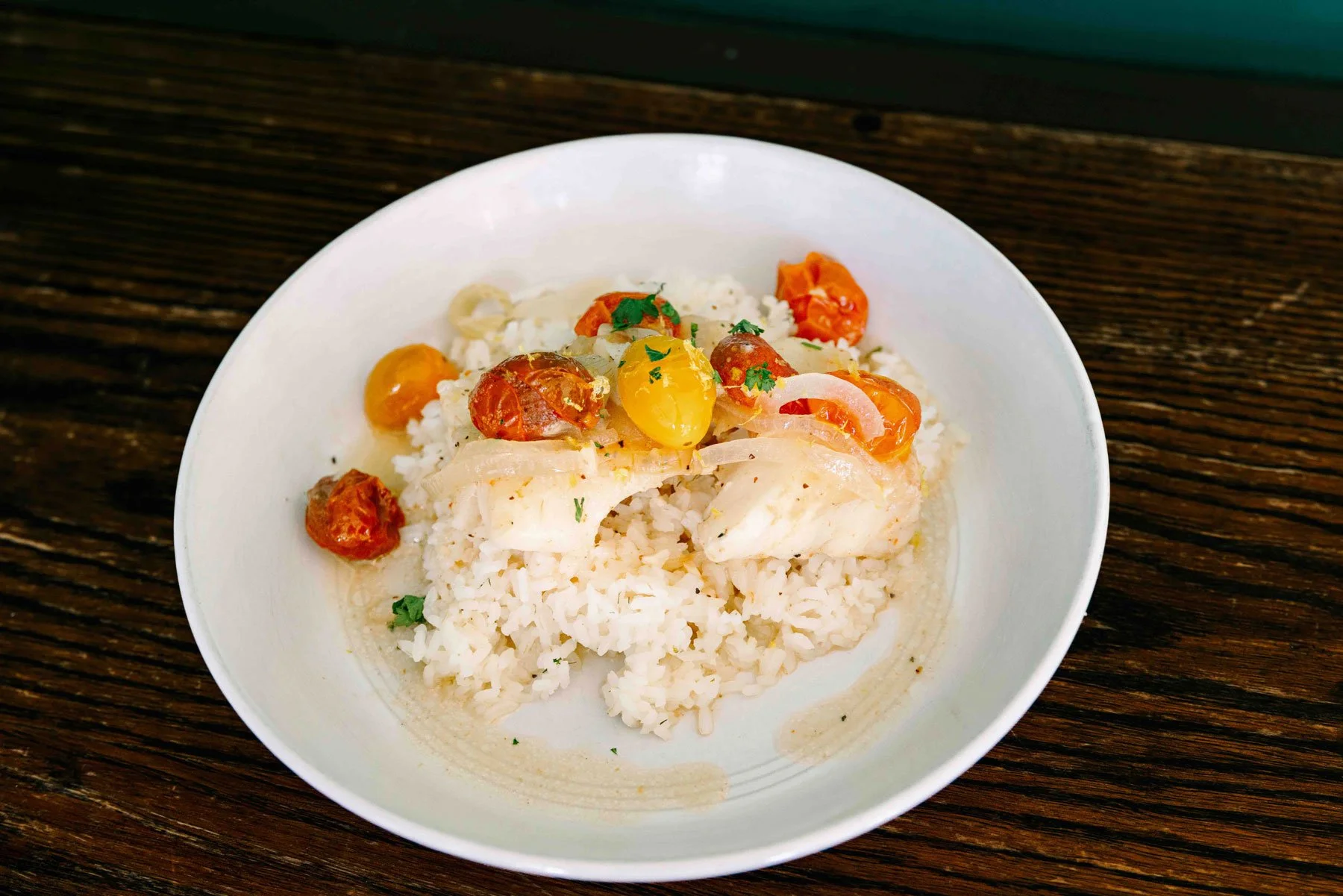 White rice topped with white fish, cherry tomatoes, and herbs on a white plate with a wooden table background.
