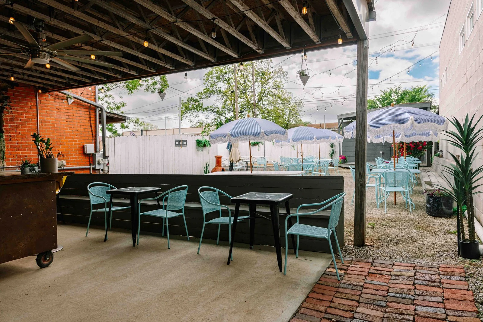 Outdoor patio area with blue chairs, tables, and umbrellas, decorated with potted plants and string lights, adjacent to a brick building and a white fence with trees in the background.