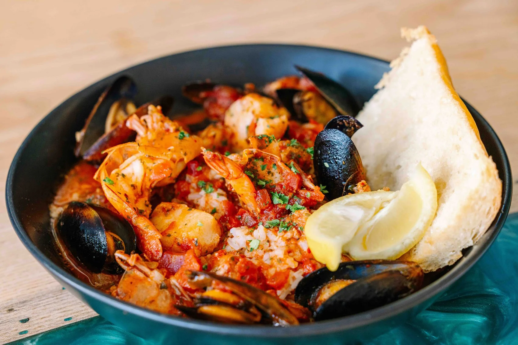 Seafood dish in a black bowl with shrimp, mussels, chopped herbs, lemon wedges, and a slice of bread.