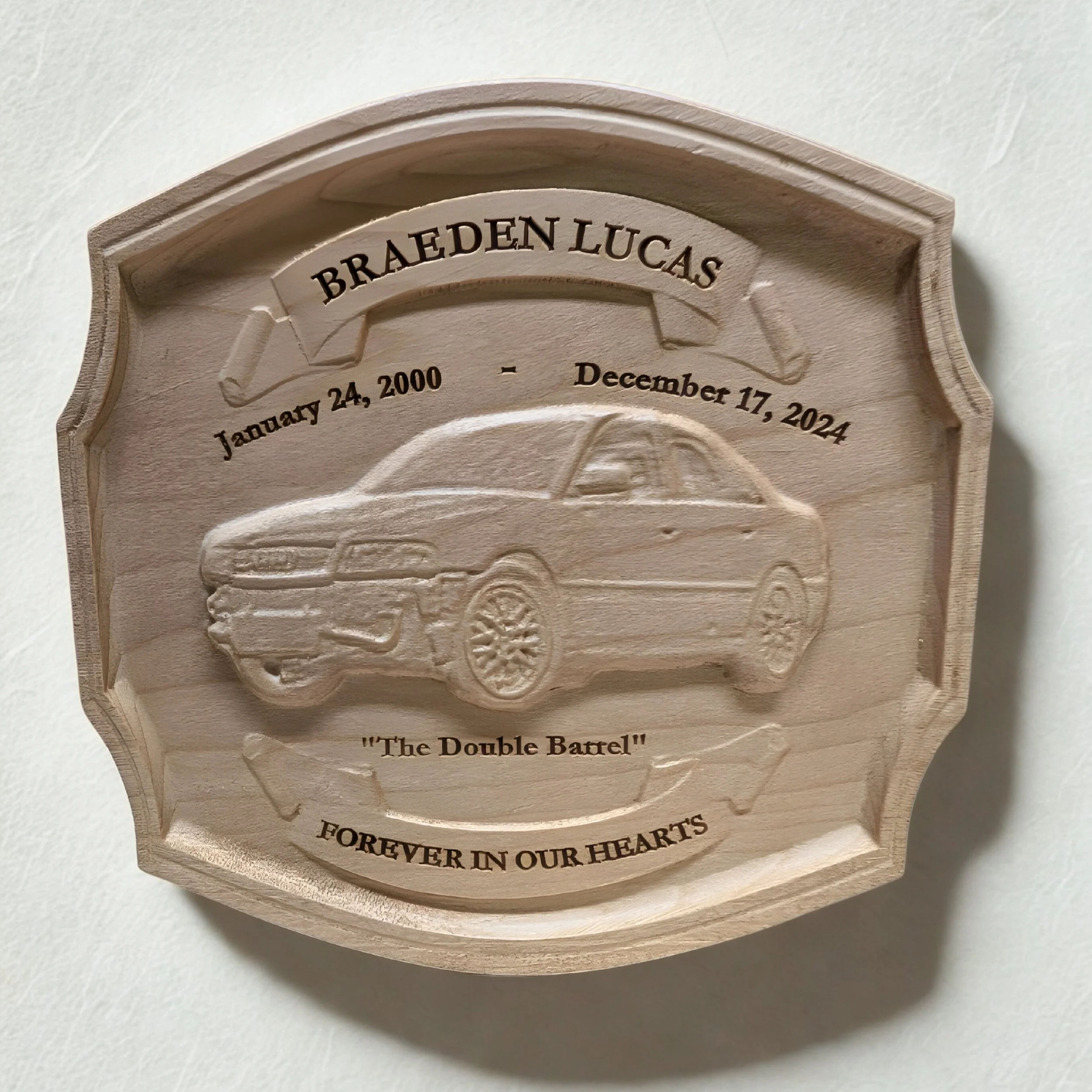 Wooden memorial plaque with a carved car in the center, inscribed with the name Braeden Lucas, birth date January 24, 2000, and death date December 17, 2024, titled 'The Double Barrel' and 'Forever in Our Hearts'.