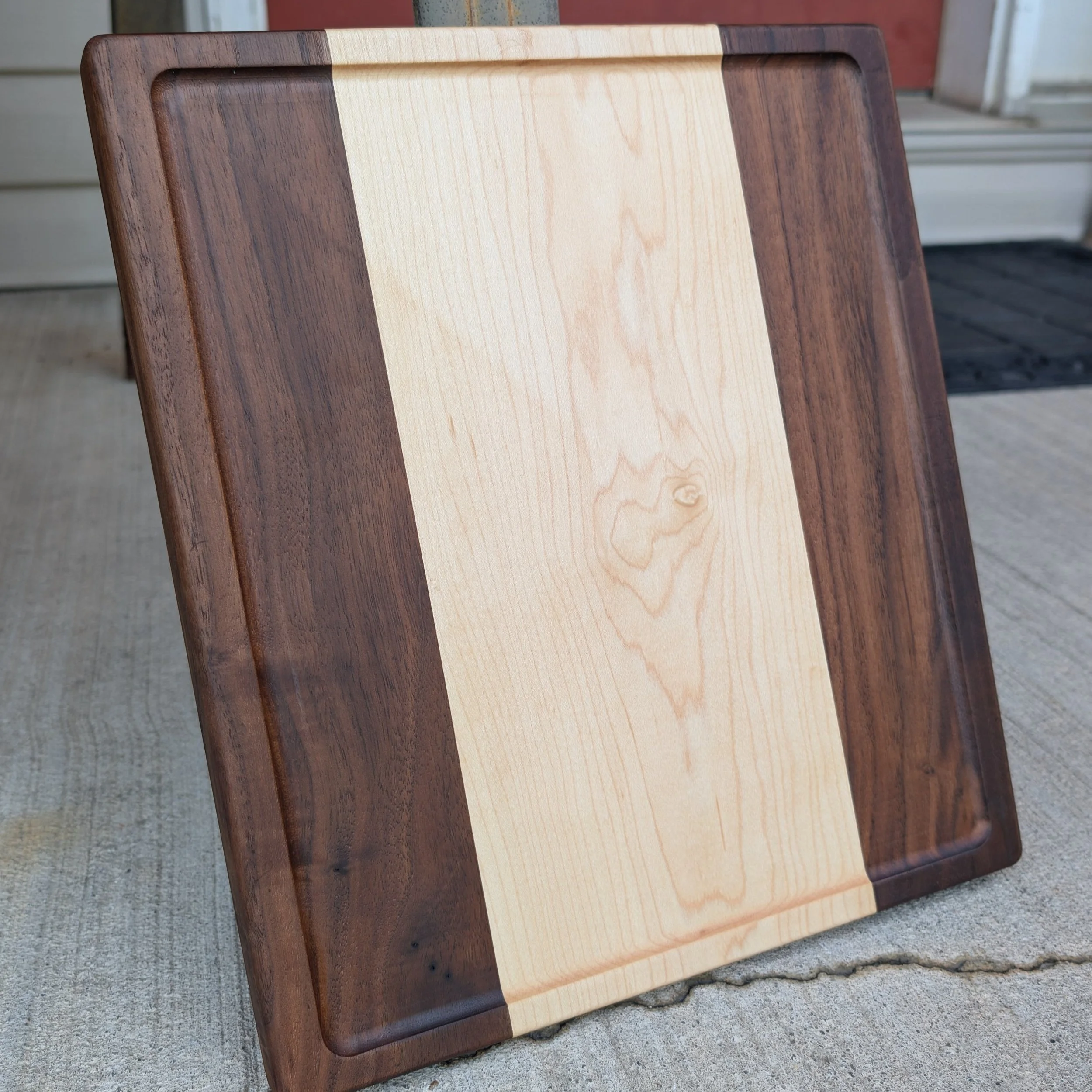 A wooden cutting board with a two-tone design, featuring a light center panel and darker border, placed on concrete outside near a house.