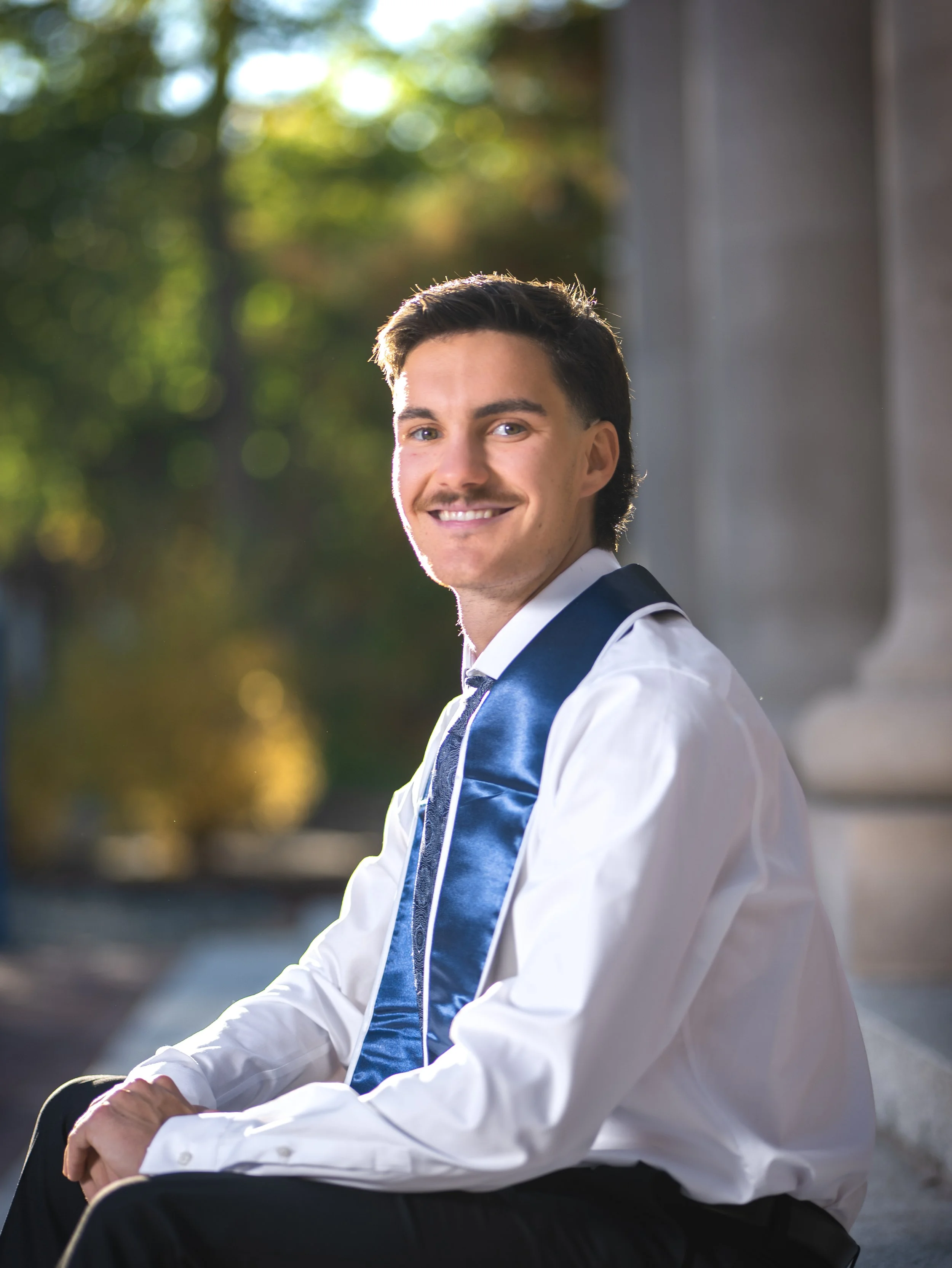 A young man in a white shirt and dark tie, wearing a blue sash, sitting outdoors on stone steps, smiling at the camera with a blurred natural background.