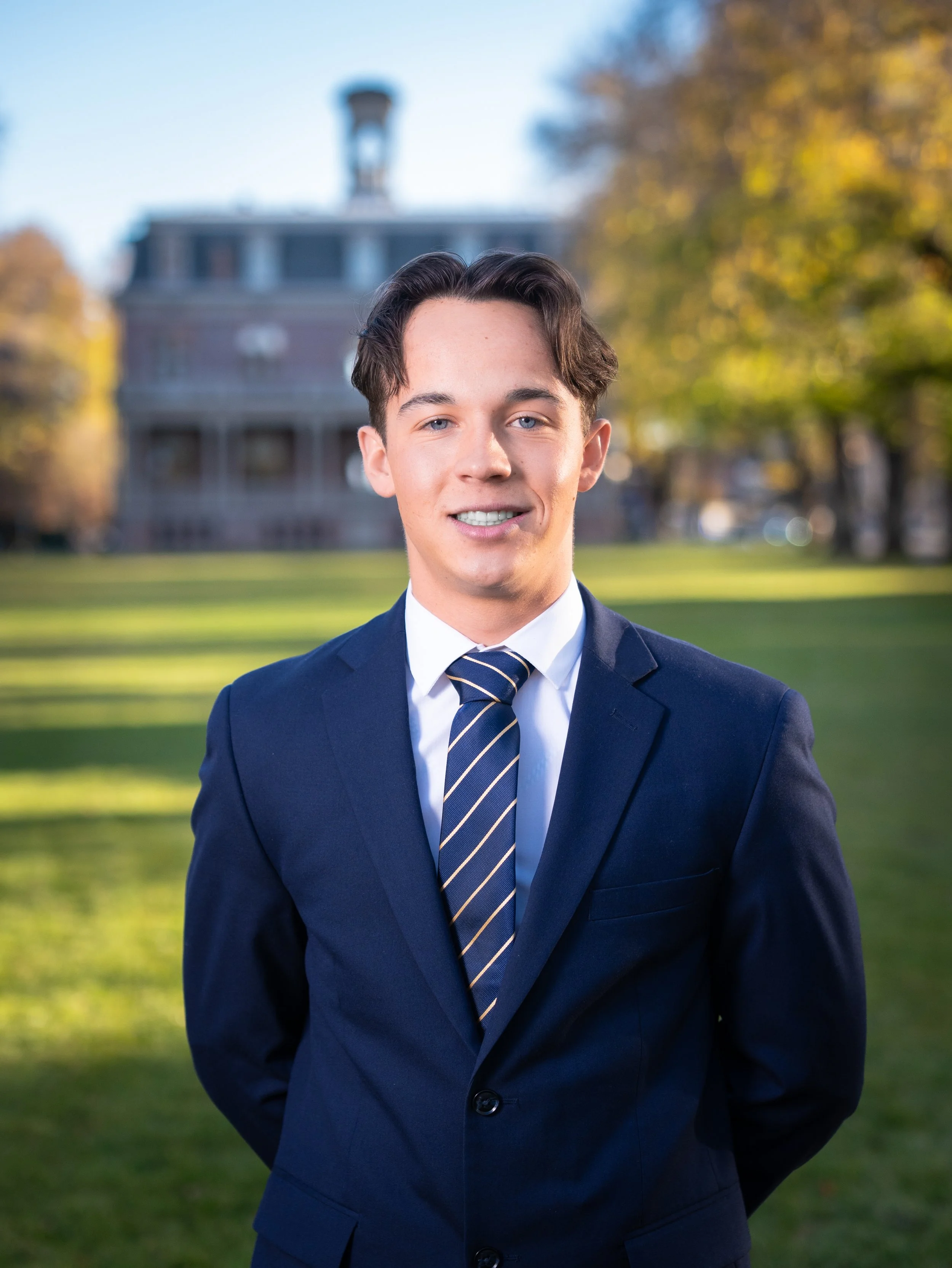 A young man in a navy suit and striped tie standing outdoors on a sunny day with a historic building and trees in the background.