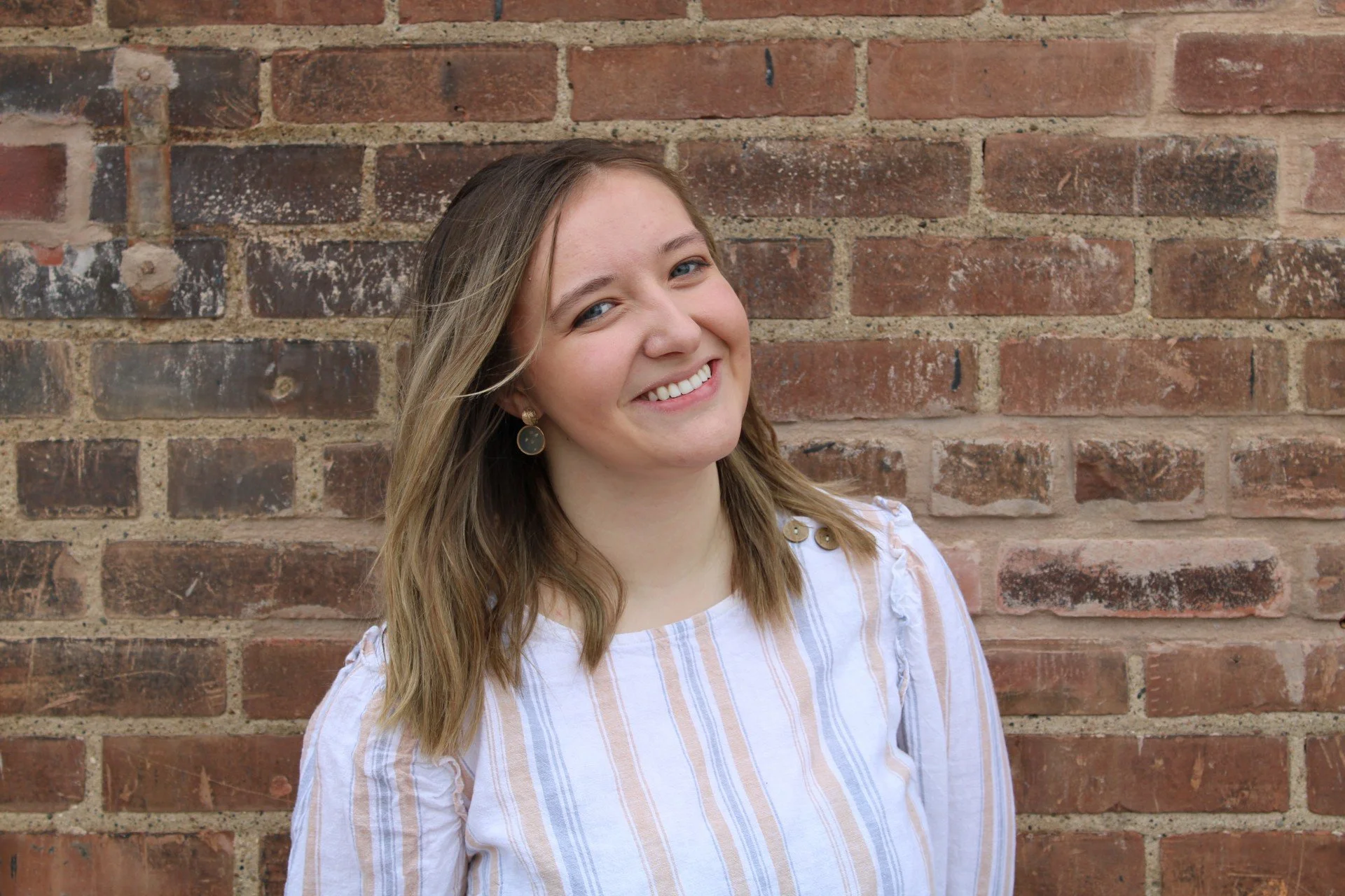 A smiling young woman with shoulder-length light brown hair, wearing earrings and a white striped shirt, stands in front of a brick wall.