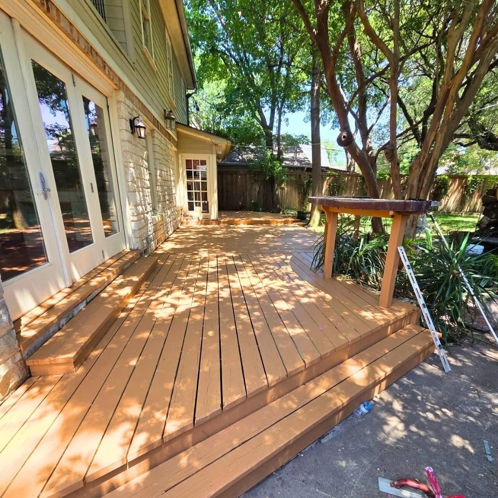 Wooden deck outside a house with folding doors, surrounded by trees and plants, partially shaded by tree shadows.
