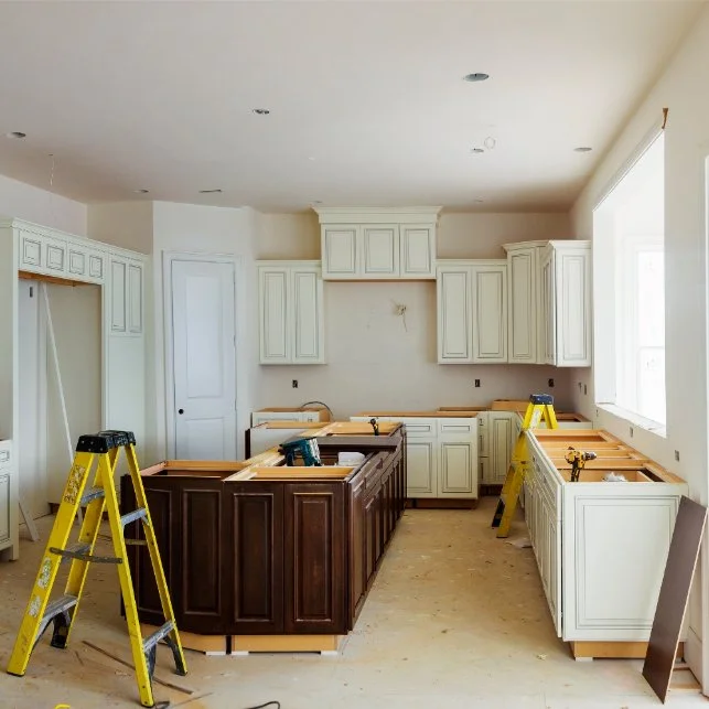 Kitchen under renovation with white and dark wood cabinetry, yellow step ladders, and construction tools.