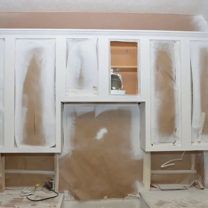 Kitchen cabinets being painted or primed, with protective paper and masking tape, over a wall with some sanding or spackling.