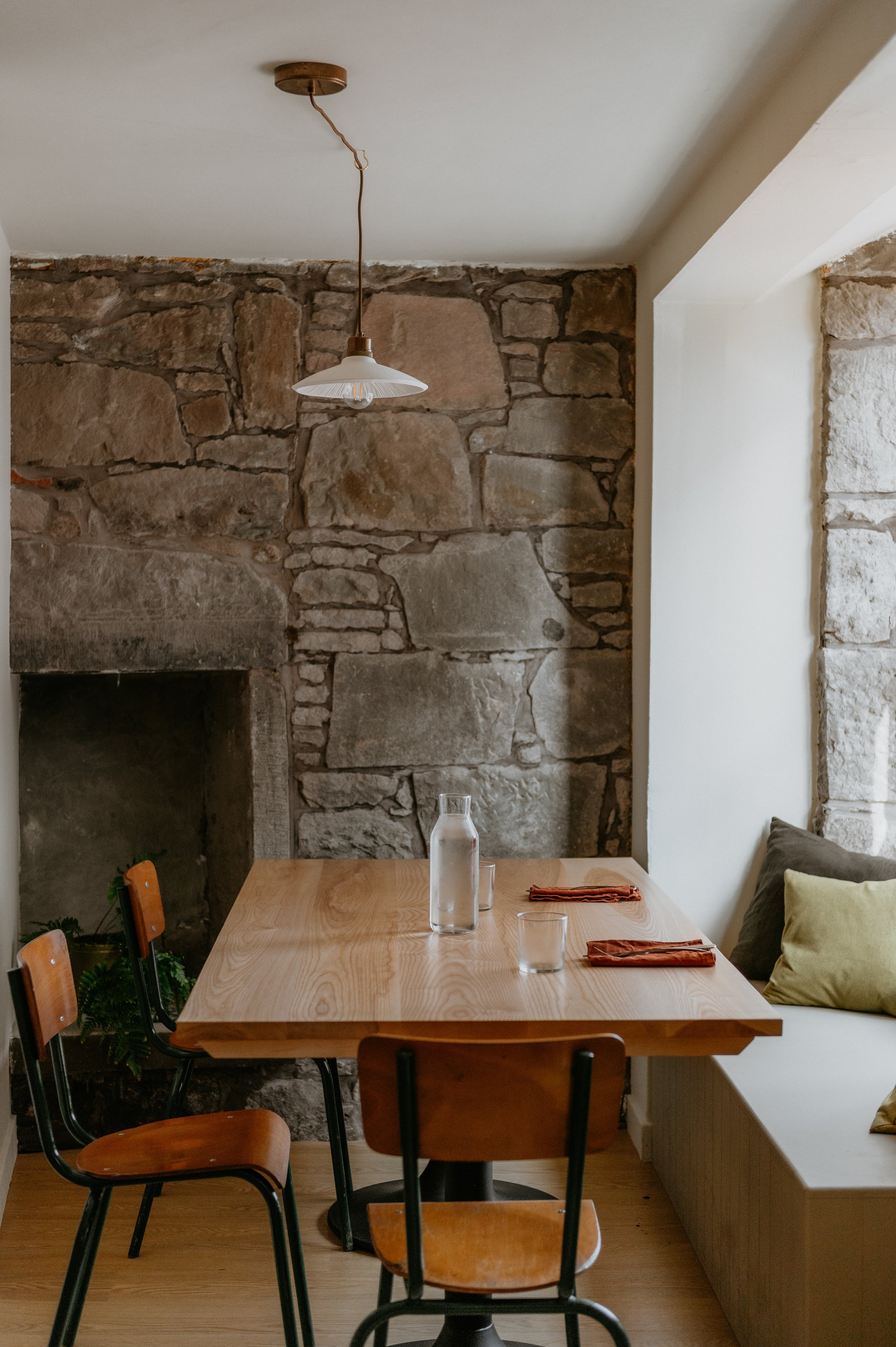A wooden dining table with a glass water bottle, two glasses, and orange napkins, set against a stone wall with a fireplace and a cushioned bench with pillows.