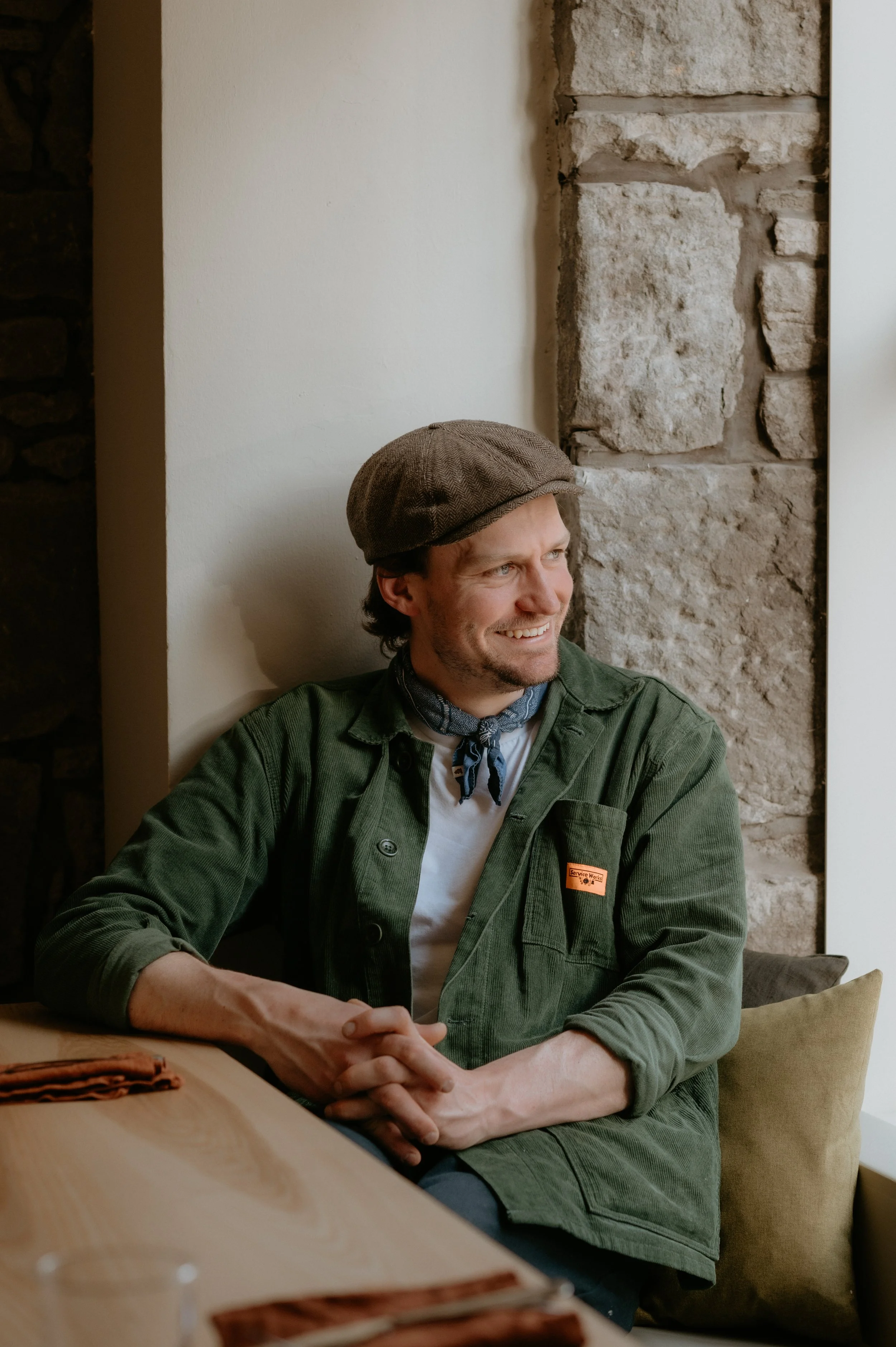 Smiling man wearing a brown hat, green jacket, and bandana sitting at a wooden table near a stone wall.
