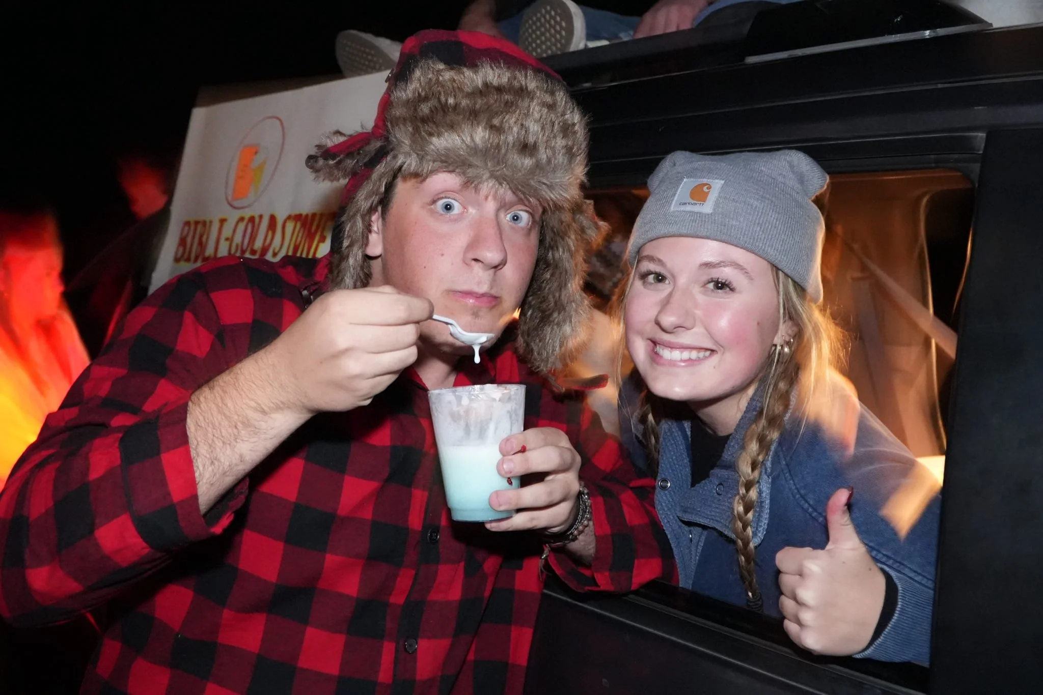Two young people with warm clothing, one wearing a furry hat, sharing a drink through a straw at a night event.