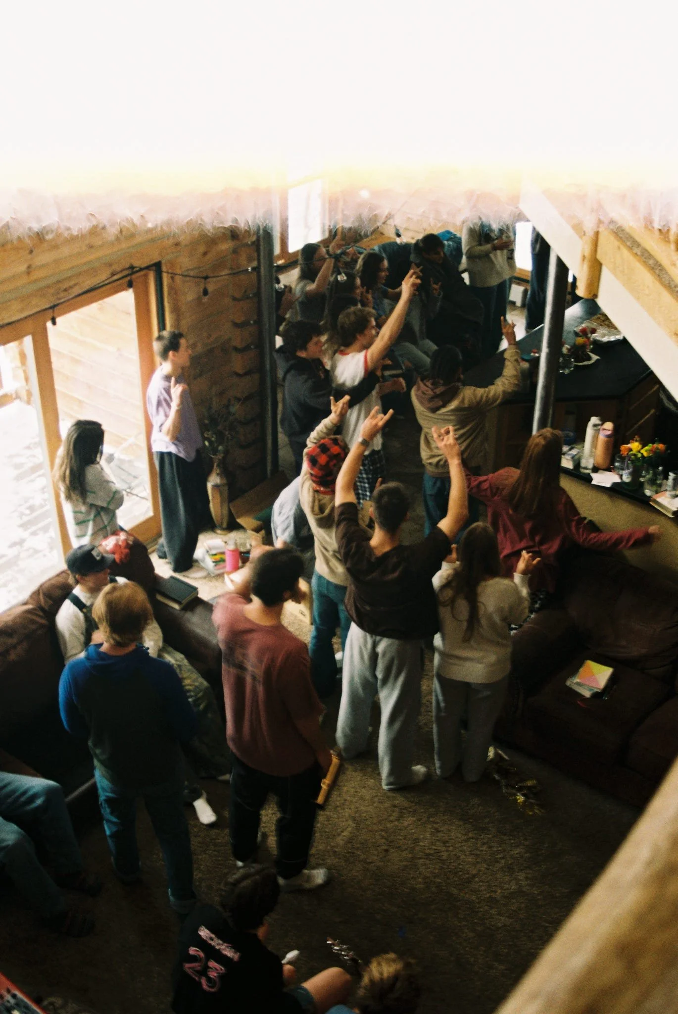 Group of people gathered in a cozy, wood-paneled room, some with hands raised, participating in a lively event near a bar area with drinks and flowers on the counter.
