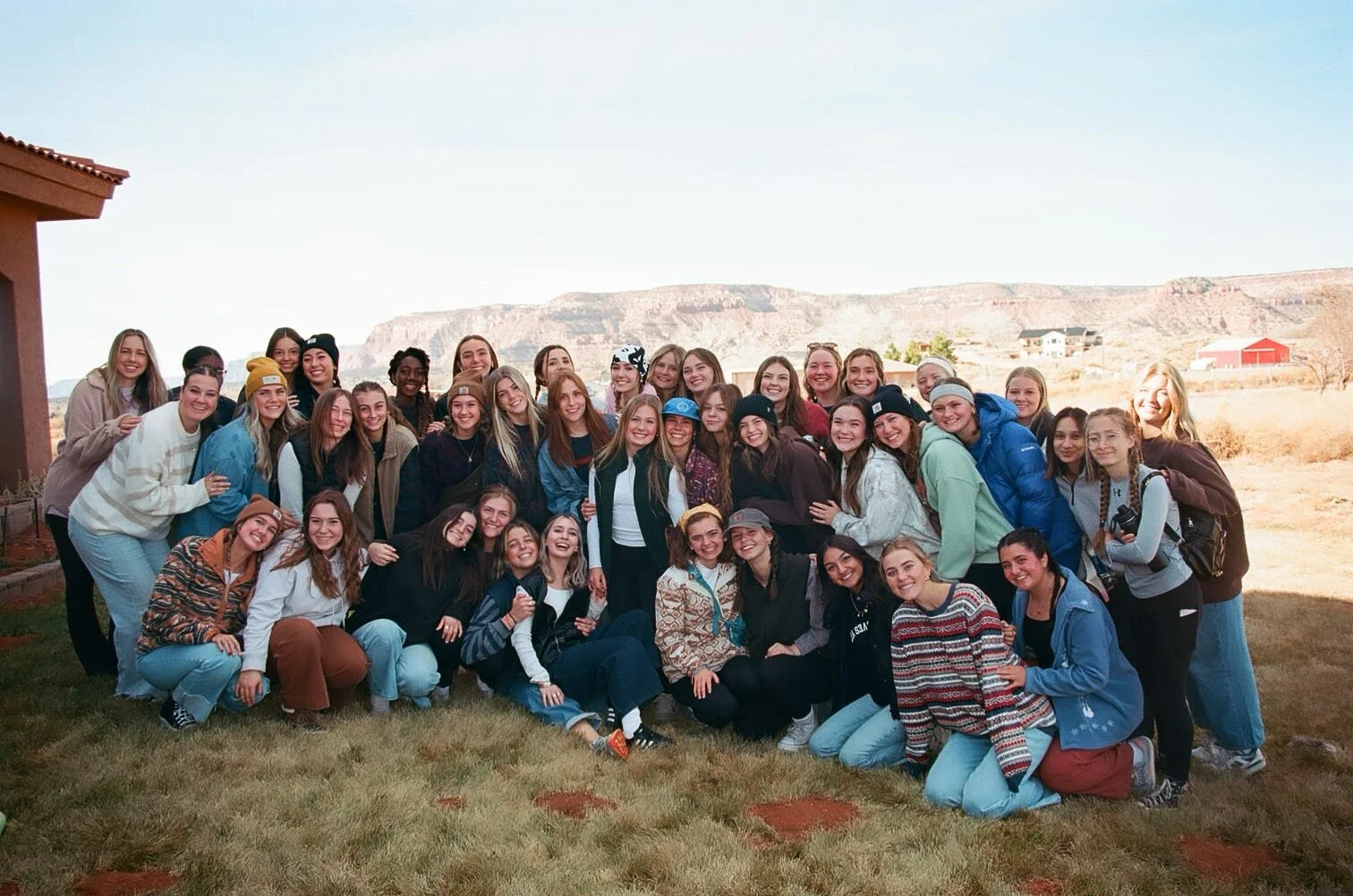 Group of smiling young women gather outdoors in a rural area with mountains and open land in the background.