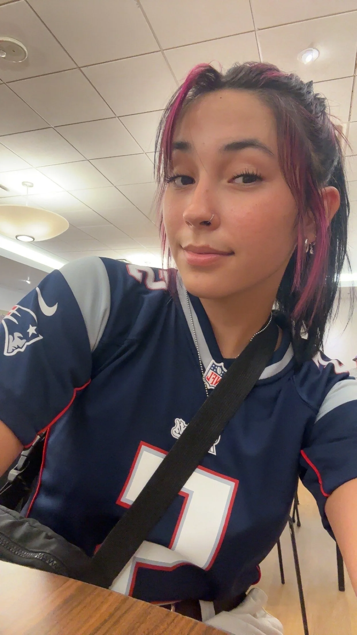 A young woman with black and pink hair, wearing a navy blue New England Patriots football jersey, sitting at a table in a well-lit indoor space.