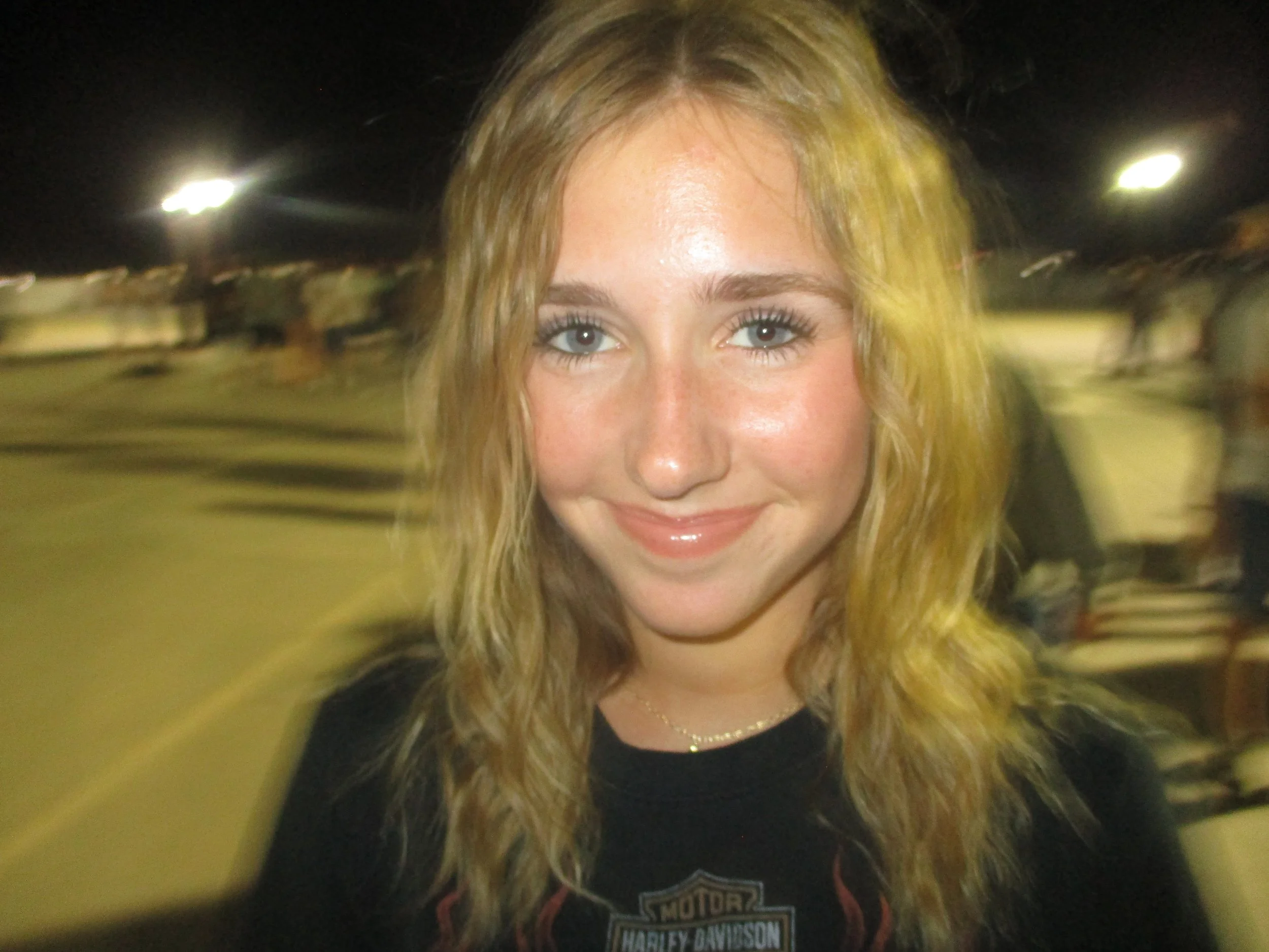 A woman with wavy blonde hair and blue eyes smiling at the camera at night, with a blurred outdoor background.