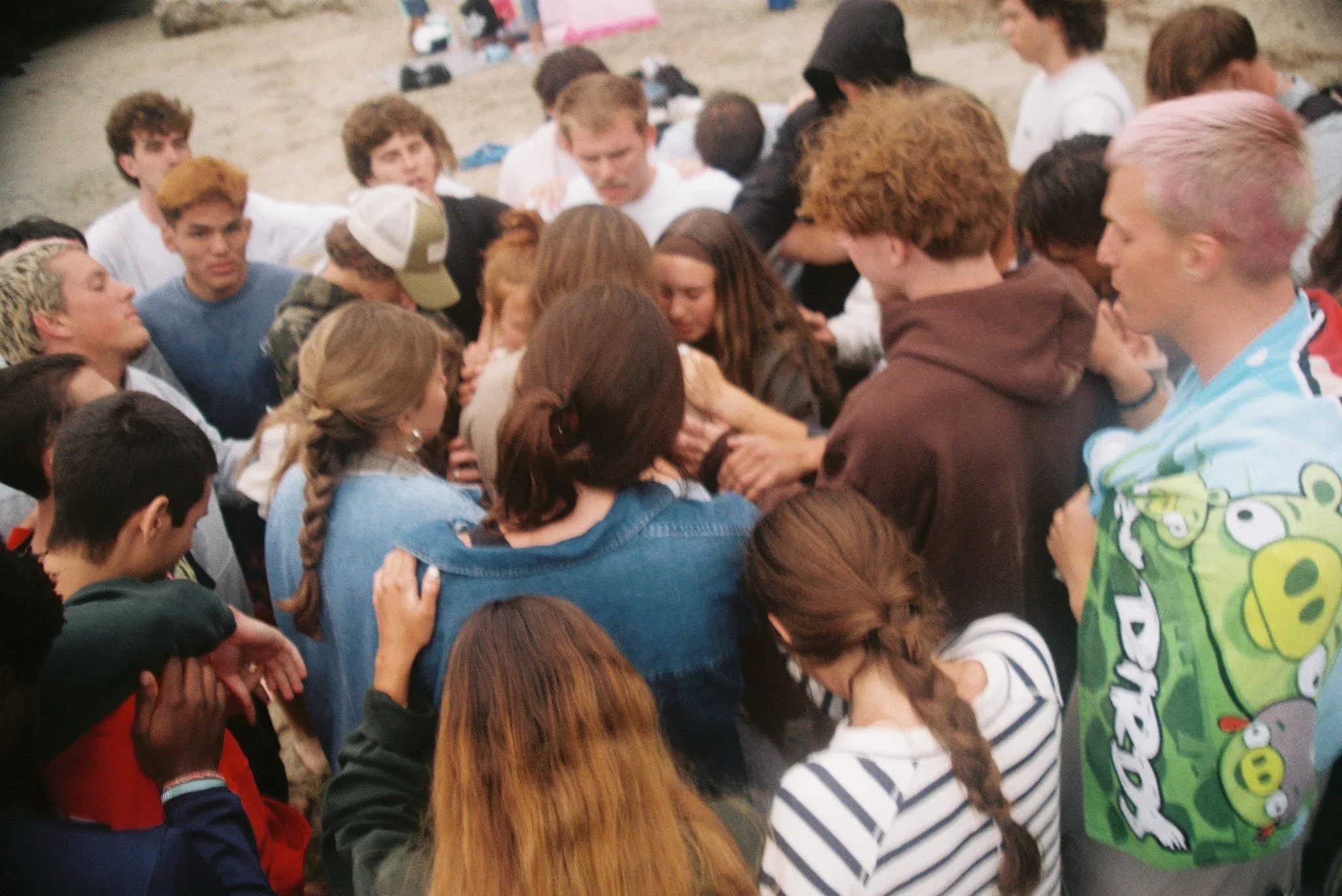 A large group of young people gathered outdoors on a beach, engaged in a moment of prayer or reflection, with some individuals holding hands or embracing.