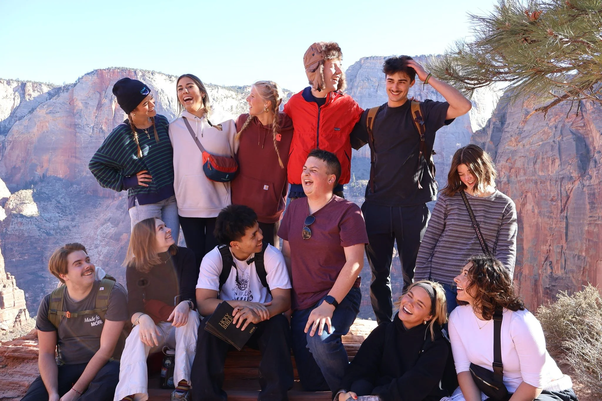 Group of friends enjoying a hike in a canyon with red rock cliffs in the background, some are sitting, some are standing, all smiling and laughing.