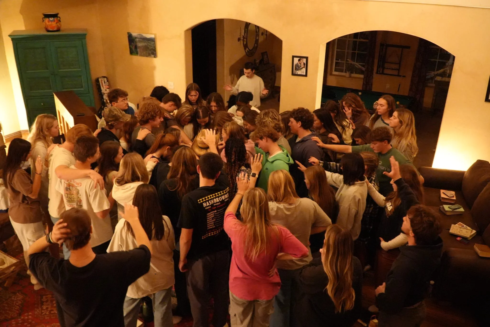 A large group of teenagers gathered closely indoors, some with arms around each other, participating in a group activity or prayer in a warmly lit living room.