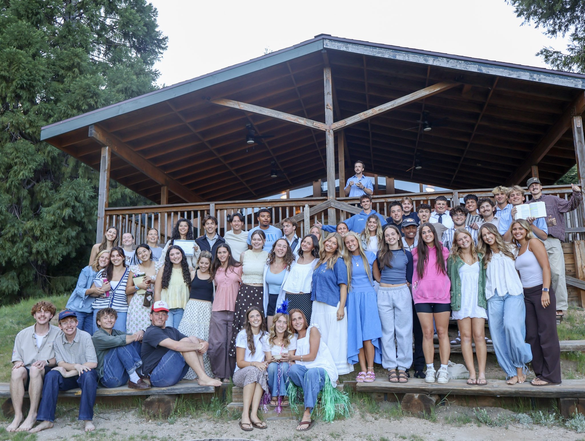 Group of young people and adults gathered outside on steps in front of a wooden building, some holding diplomas, celebrating graduation or recognition event.
