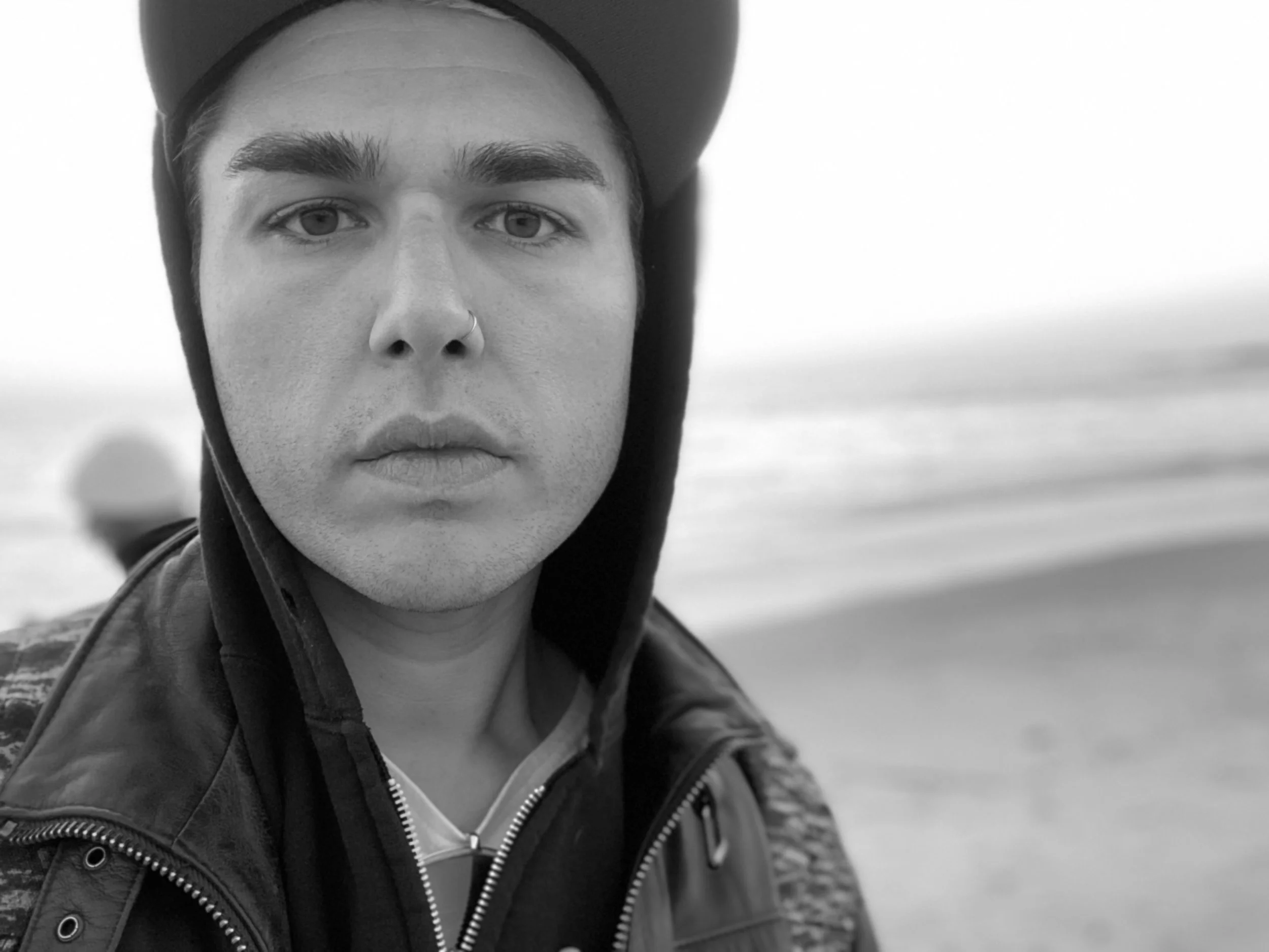 A black-and-white close-up of a young man with light skin wearing a hoodie, leather jacket, and a hat at the beach.